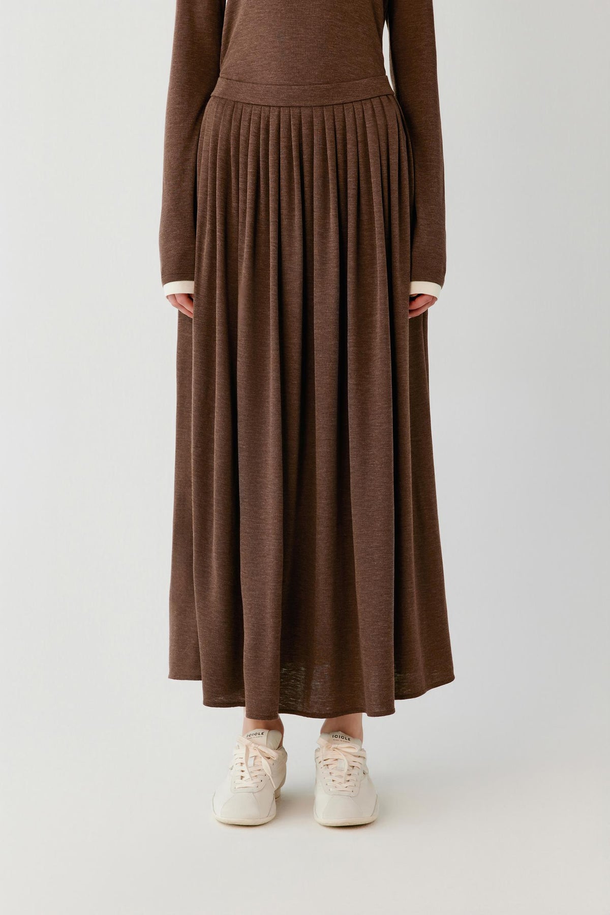 A person wearing a long-sleeve brown knit maxi dress with a pleated skirt and white sneakers, photographed against a plain light gray studio background.