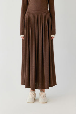 A person wearing a long-sleeve brown knit maxi dress with a pleated skirt and white sneakers, photographed against a plain light gray studio background.