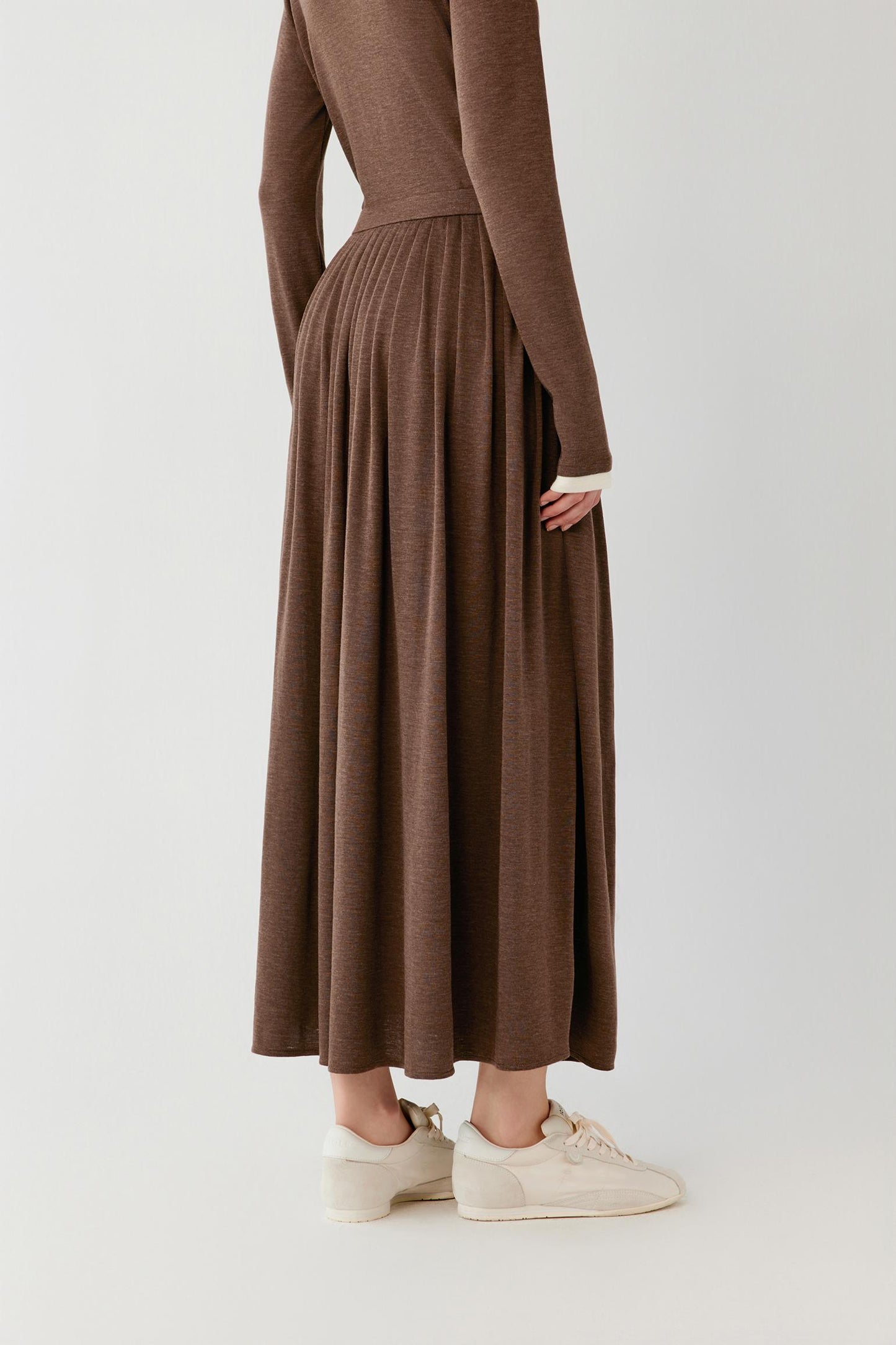 A person shown from the back wearing a long-sleeve brown knit maxi dress with pleated detailing, paired with cream sneakers, against a plain light gray studio background.