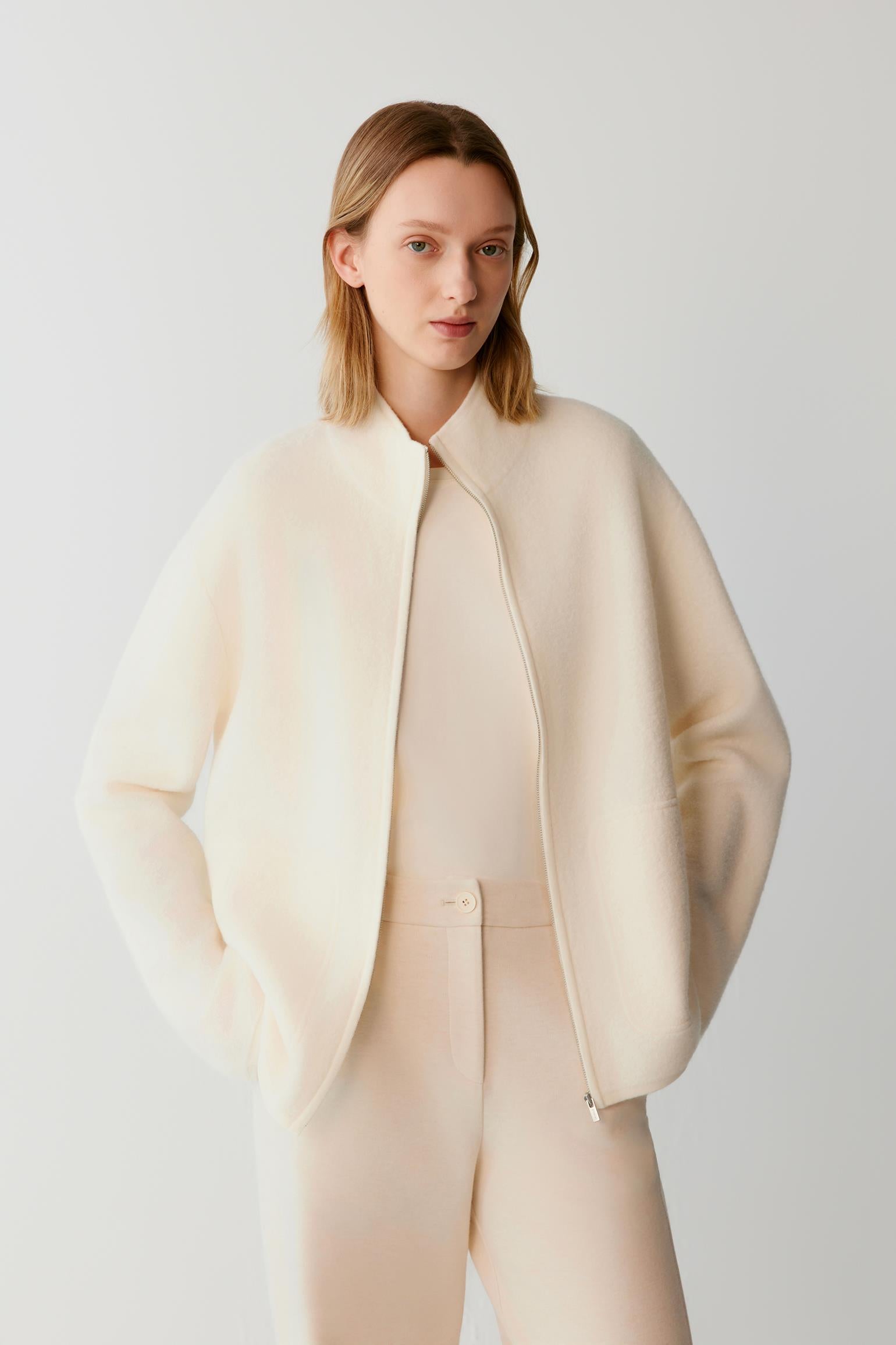 A woman modeling a cream-colored, lightweight structured jacket with a high collar and front zipper, paired with matching cream trousers, against a light gray studio background.