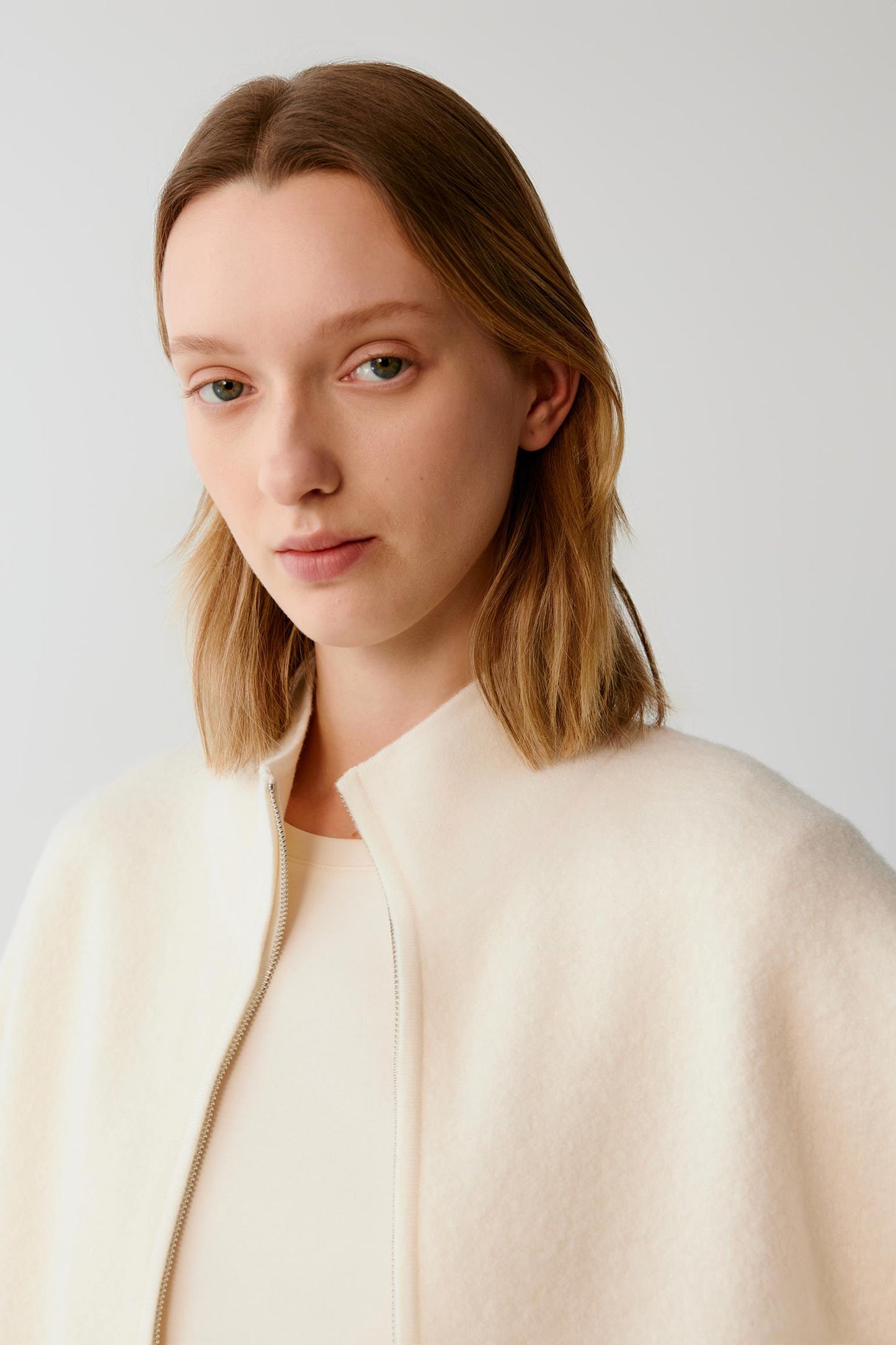 A woman with shoulder-length light brown hair wearing a cream-colored textured jacket over a matching cream top, photographed against a plain light gray studio background.