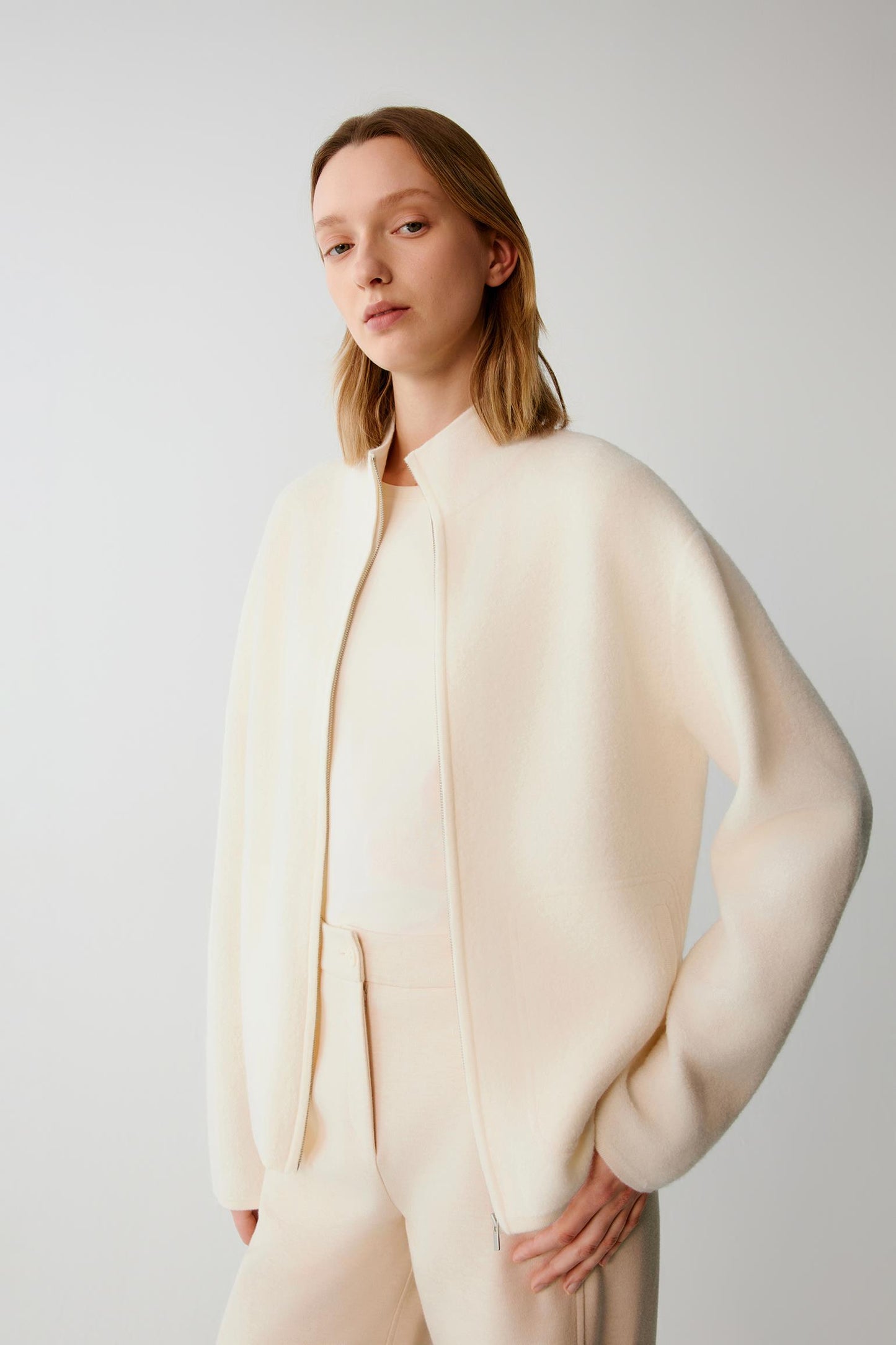 A model wearing a cream-colored, oversized knit jacket with a mock neck and long sleeves, paired with matching knit trousers, against a plain light gray studio background.