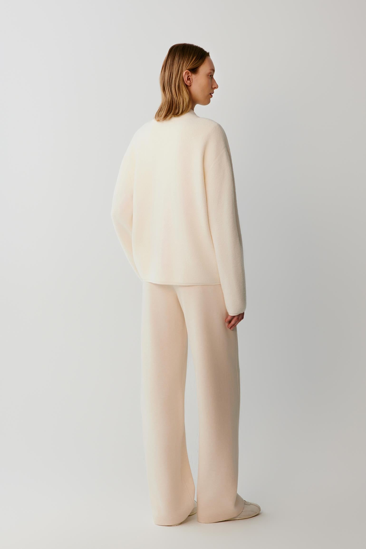 A model shown from the back wearing a matching cream-colored knit sweater and wide-leg trousers set against a plain light gray studio background.