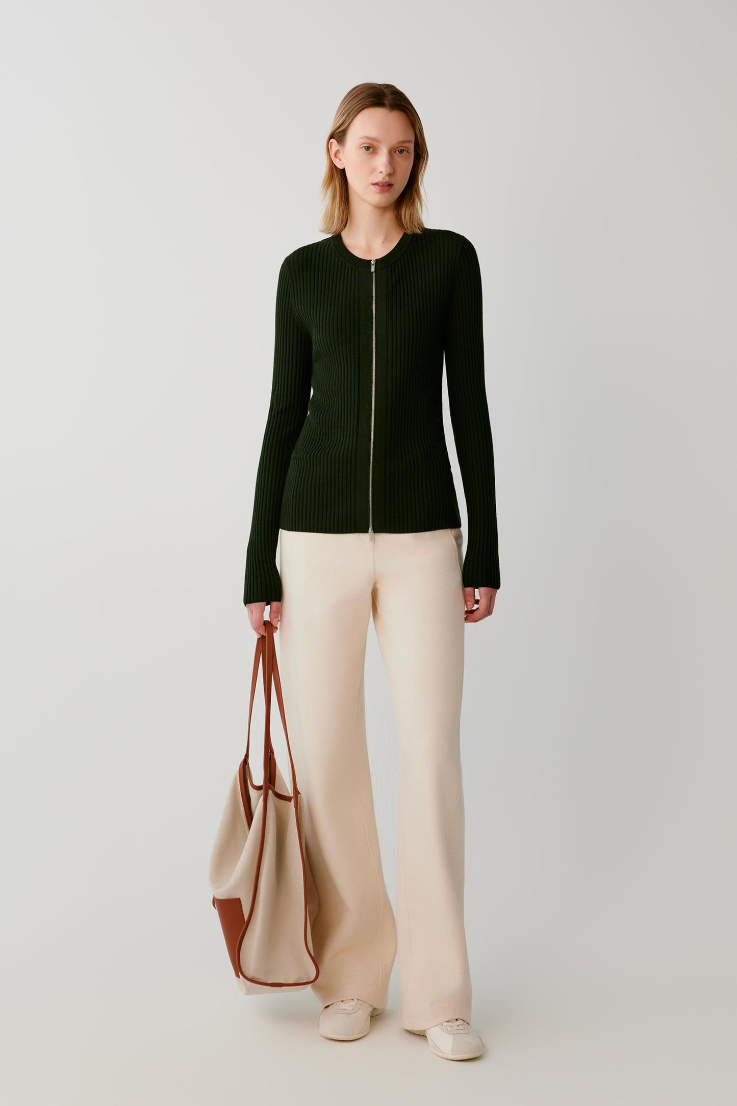 A woman stands against a light gray studio background wearing a dark green ribbed zip-front sweater, cream wide-leg pants, and holding a beige tote bag with brown handles.