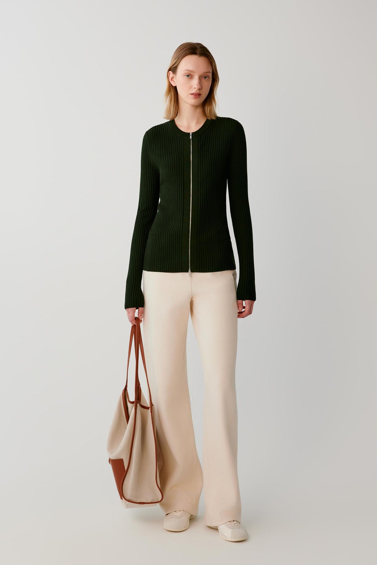 A woman stands against a light gray studio background wearing a dark green ribbed zip-front sweater, cream wide-leg pants, and holding a beige tote bag with brown handles.