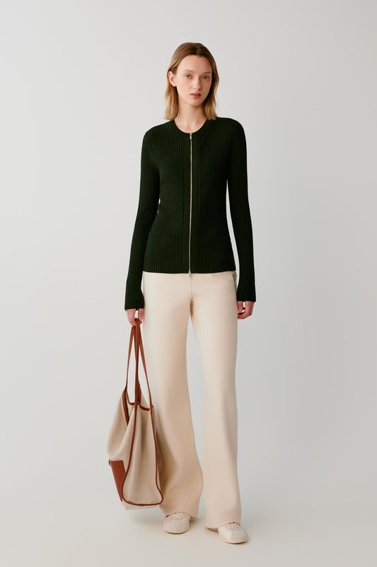 A woman stands against a light gray studio background wearing a dark green ribbed zip-front sweater, cream wide-leg pants, and holding a beige tote bag with brown handles.