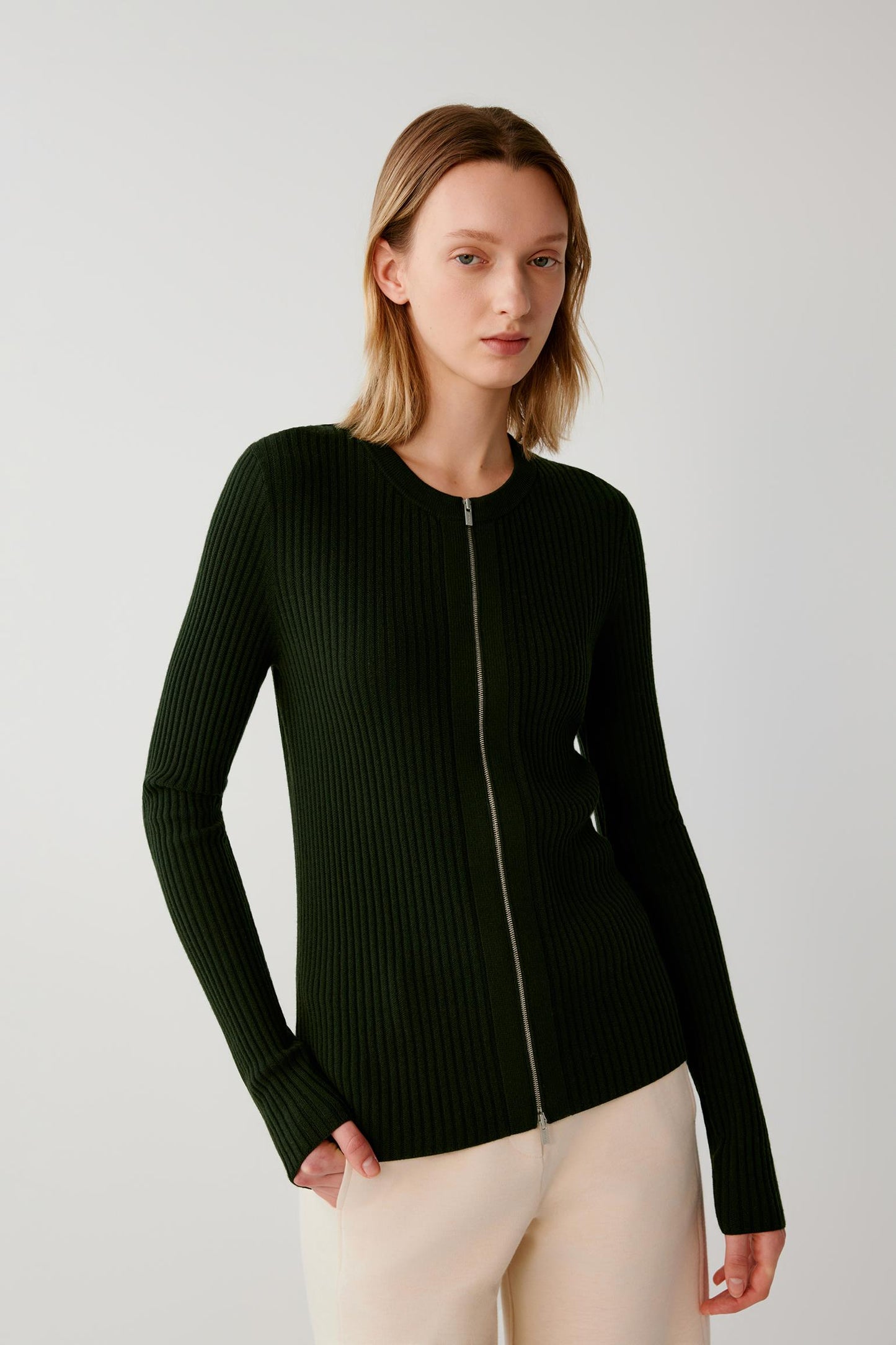 A woman modeling a dark forest green ribbed knit zip-up cardigan sweater with cream pants, photographed against a neutral light gray studio background.