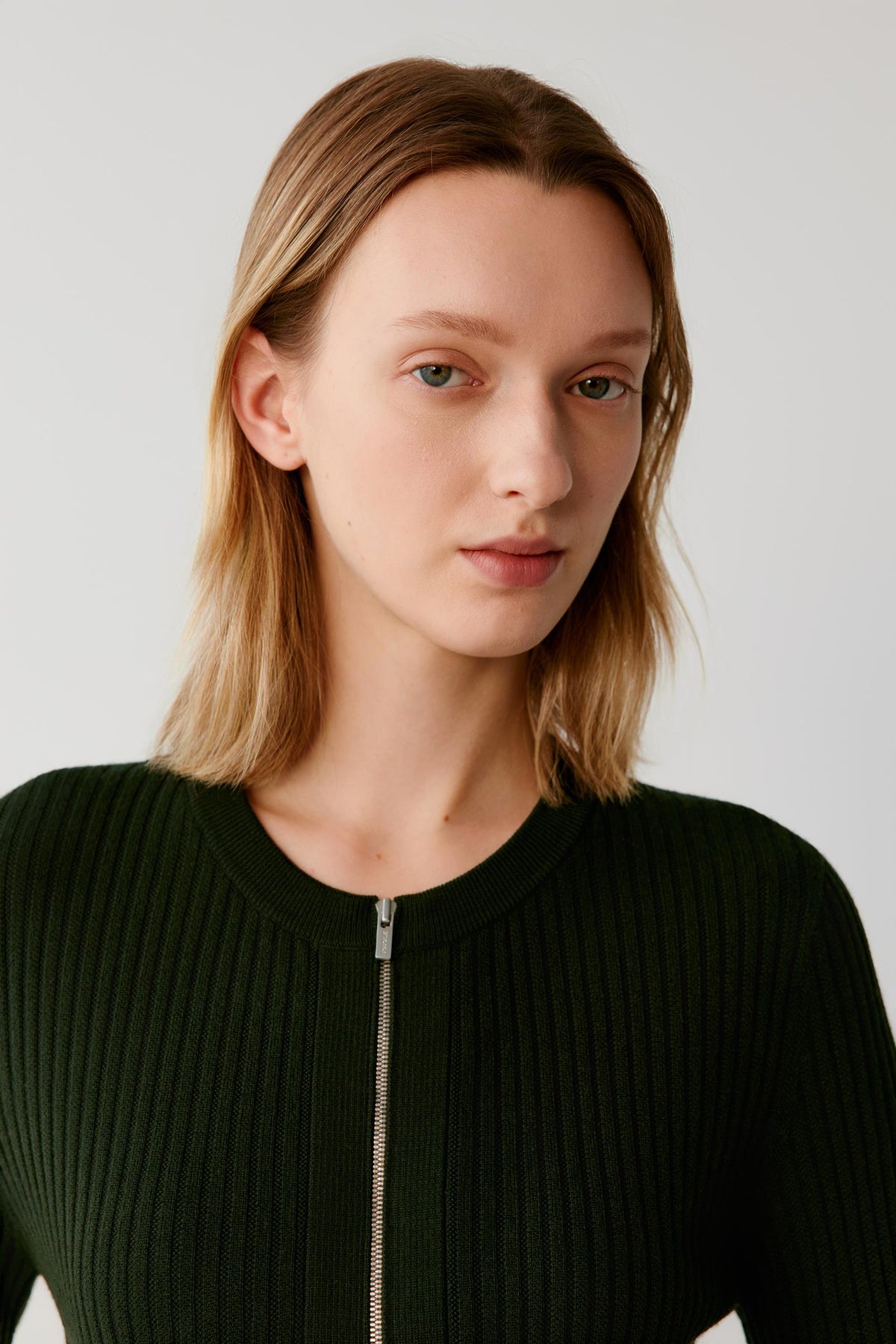 A woman with shoulder-length blonde hair wearing a dark green ribbed knit top with a silver zipper at the neckline, photographed against a light gray studio background.