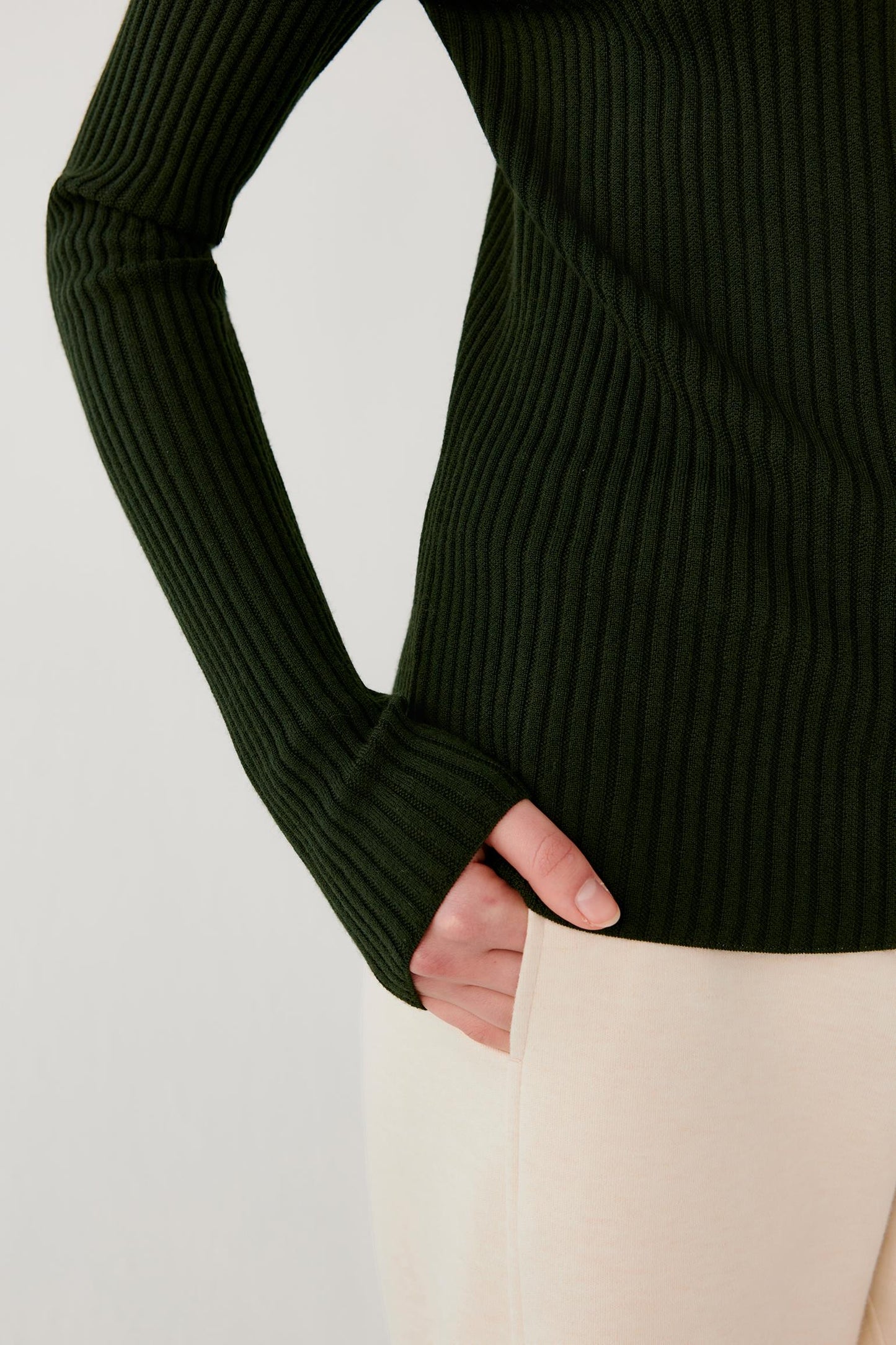 Close-up of a person wearing a dark forest green ribbed knit long-sleeve top with cream-colored pants, with one hand in a pocket.