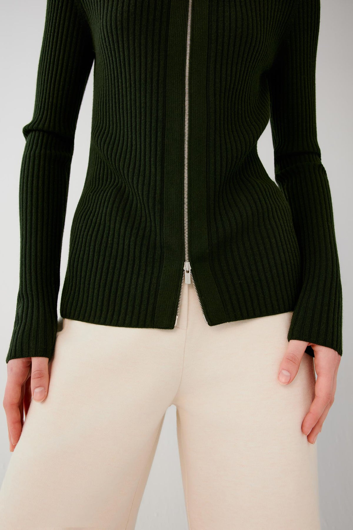 A close-up of a model wearing a dark forest green ribbed knit zip-up sweater and cream-colored pants, showing the front of the garment with a silver zipper.