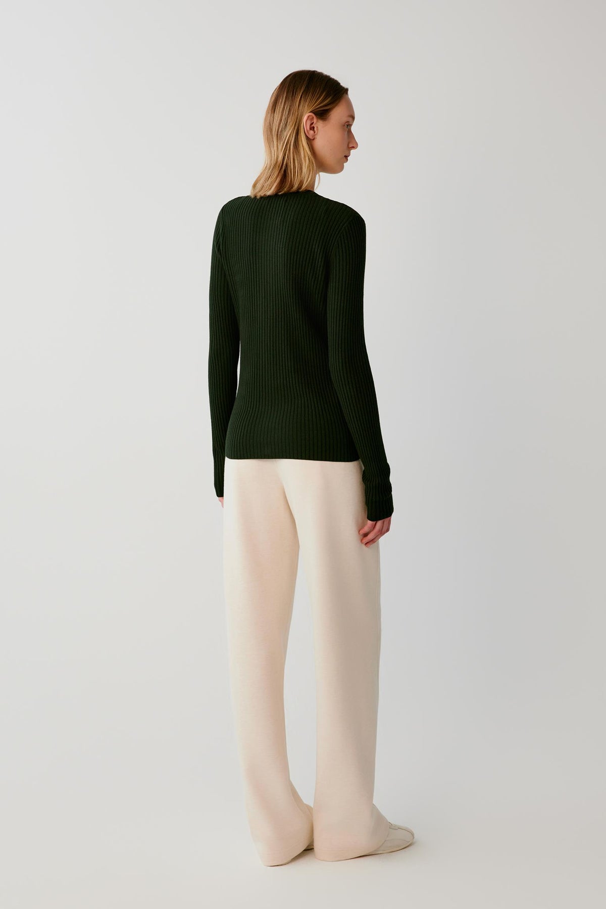 A woman stands with her back to the camera wearing a dark forest green ribbed knit sweater and cream wide-leg trousers against a plain light gray studio background.