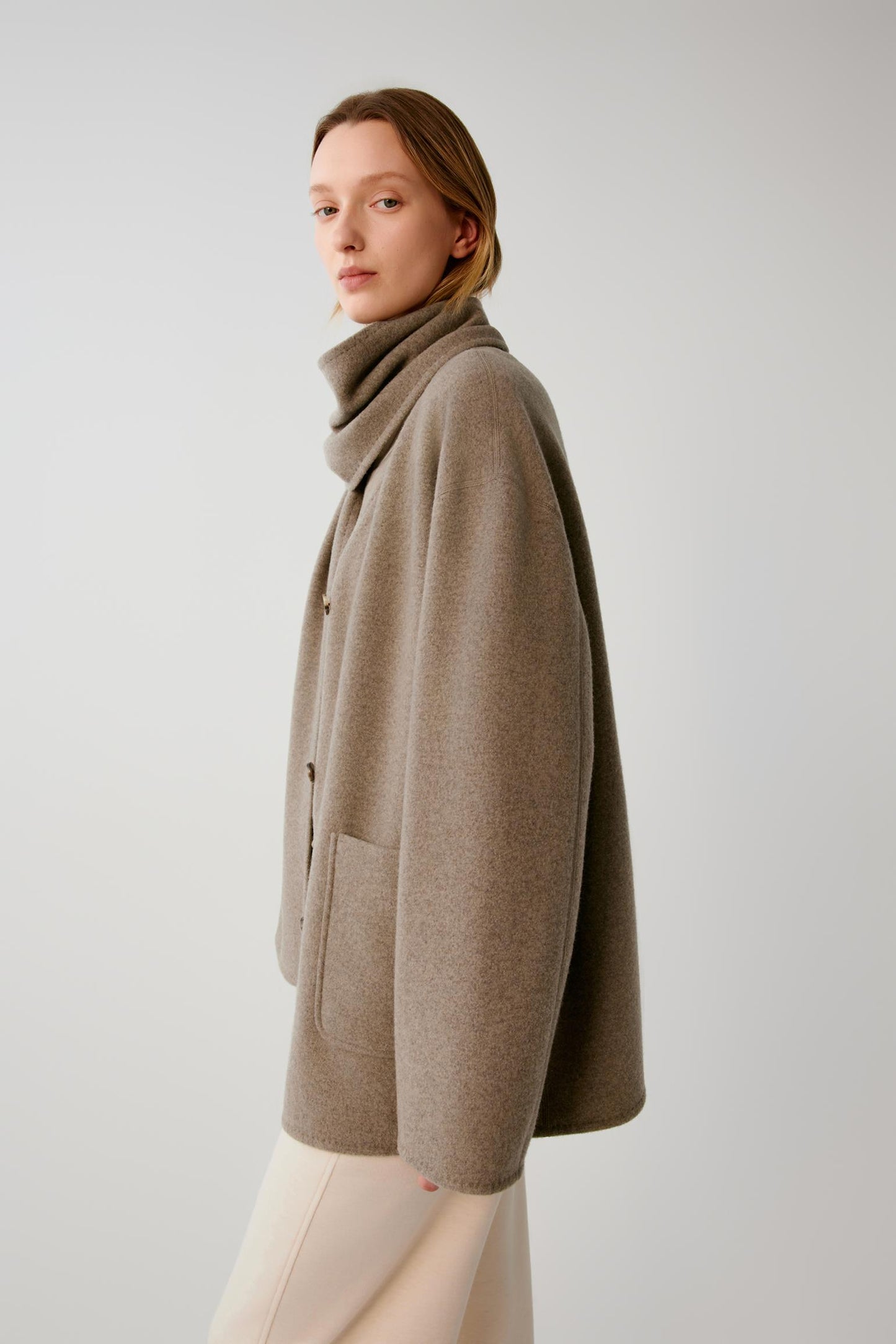 A woman wearing a taupe-brown cowl-neck cardigan with front buttons and side pockets, paired with light beige trousers, photographed against a neutral light gray studio background.