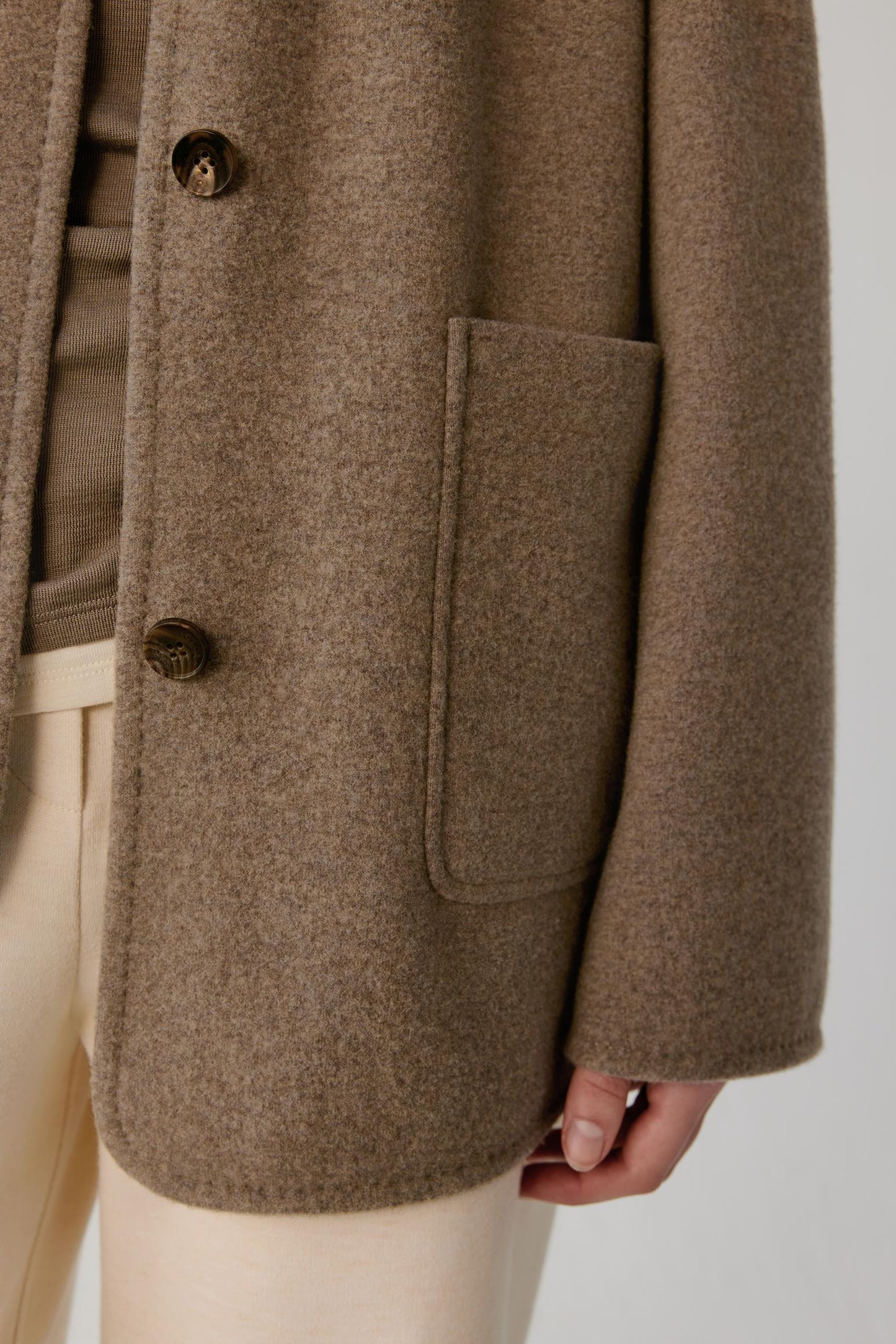 Close-up of a taupe-brown wool coat with dark brown buttons and a front patch pocket, worn over a beige top and cream trousers.