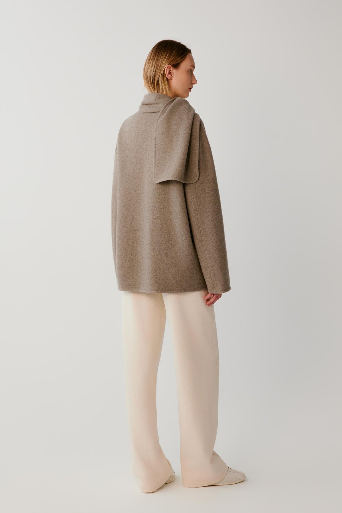 A model shown from the back wearing a taupe-brown oversized knit wrap coat and cream wide-leg trousers, standing against a light gray studio background.
