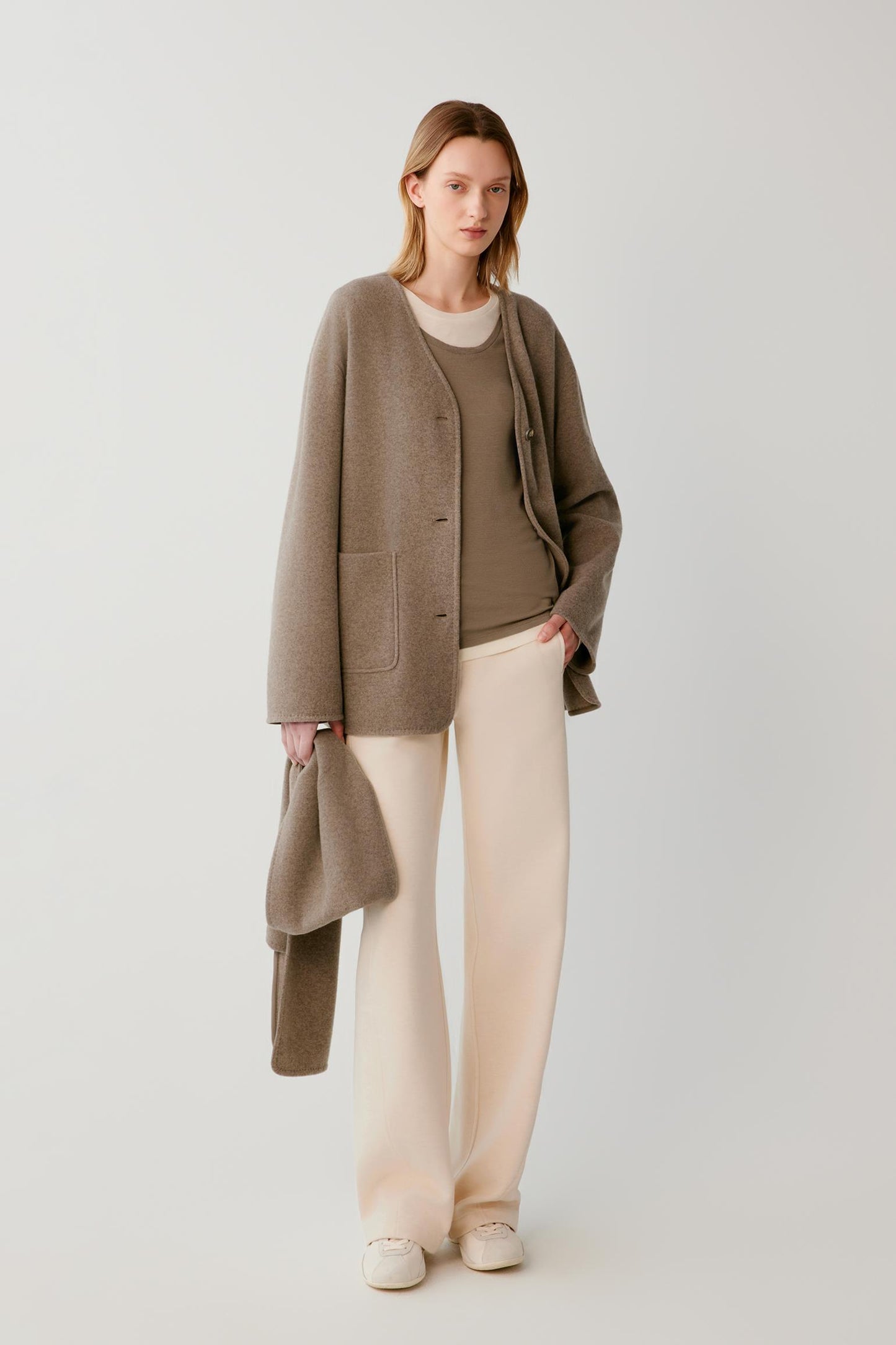 A woman stands against a light gray studio background wearing a taupe open-front cardigan over a matching taupe top, cream wide-leg pants, and cream shoes, while holding a taupe clutch.