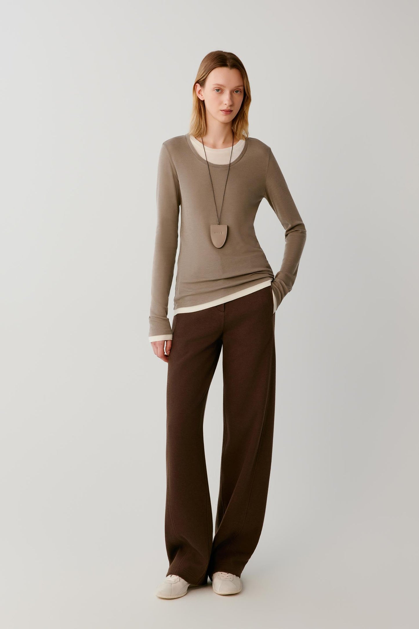 A model stands against a light gray studio background wearing a taupe long-sleeve knit top over a white undershirt, dark brown wide-leg trousers, and off-white slip-on shoes.