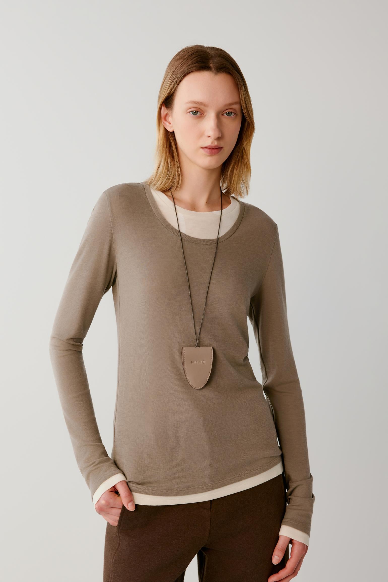 A woman modeling a taupe long-sleeve crewneck sweater with contrasting cream ribbed trim at the collar, hem, and cuffs, paired with dark brown trousers and a shield-shaped pendant necklace, against a light gray studio background.