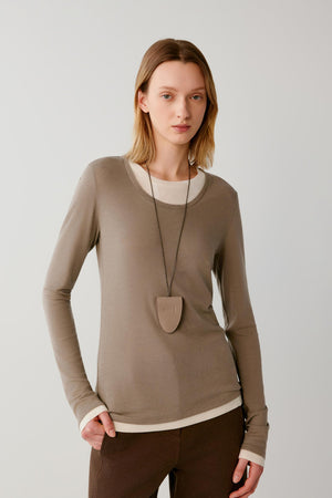 A woman modeling a taupe long-sleeve crewneck sweater with contrasting cream ribbed trim at the collar, hem, and cuffs, paired with dark brown trousers and a shield-shaped pendant necklace, against a light gray studio background.
