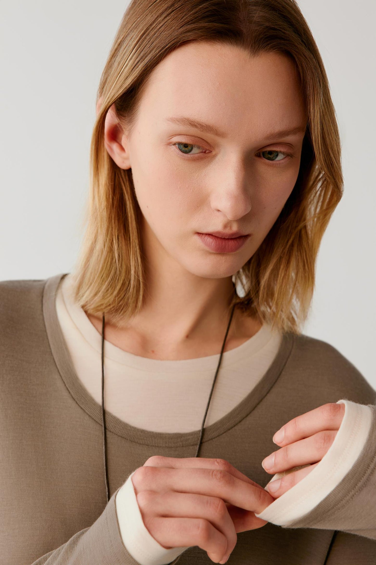 A close-up of a woman wearing a layered knit top with a taupe-brown outer layer and a cream inner layer, paired with a black cord necklace.