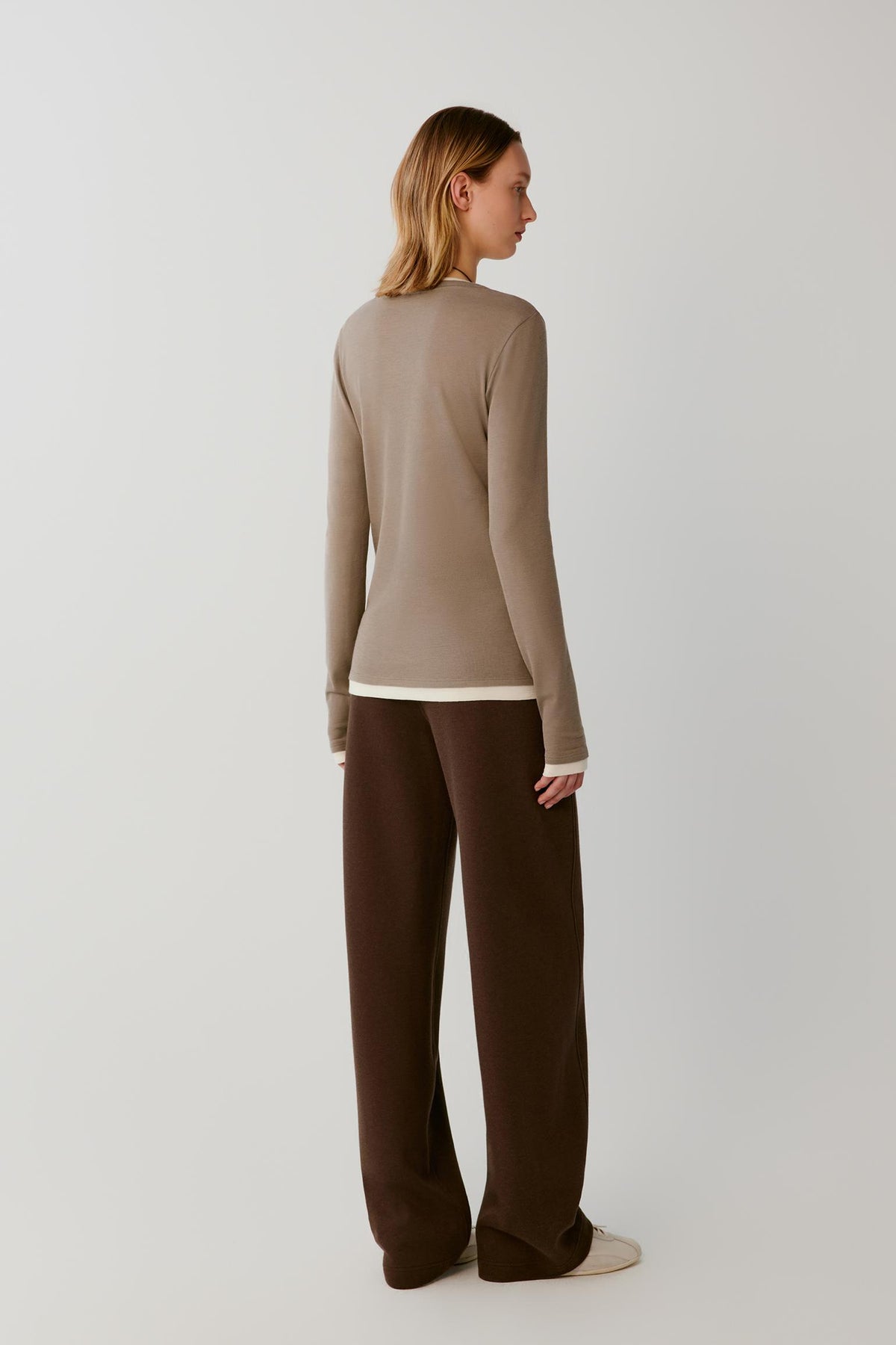 A woman shown from the back wearing a beige long-sleeve top and dark brown wide-leg trousers, standing against a plain light gray studio background.