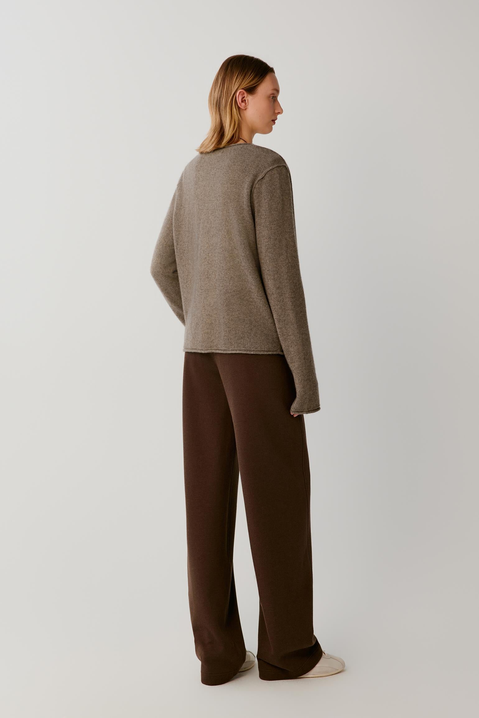 A woman stands in profile wearing a taupe knit sweater and dark brown wide-leg trousers against a plain light gray studio background.
