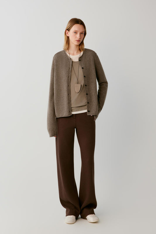 A model stands against a light gray studio background wearing a taupe knit cardigan over a beige top, dark brown wide-leg trousers, and off-white slip-on shoes.