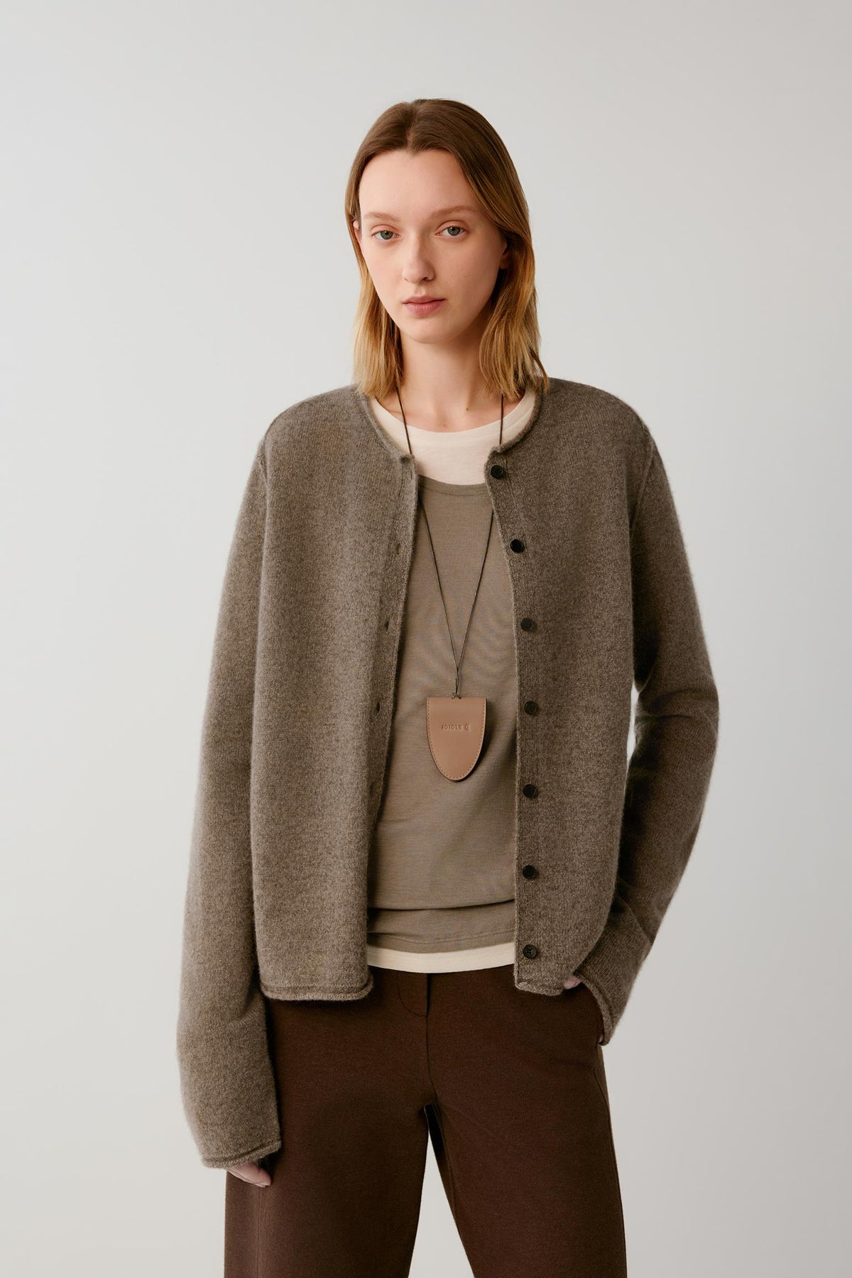 A woman modeling a taupe brown button-up cardigan over a beige top and brown trousers, set against a plain light gray studio background.