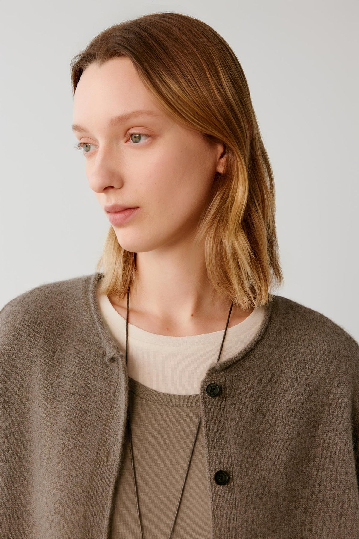 A woman with shoulder-length light brown hair is shown in a close-up portrait wearing a taupe-brown button-front cardigan over a cream top and olive green undershirt, set against a plain light gray background.