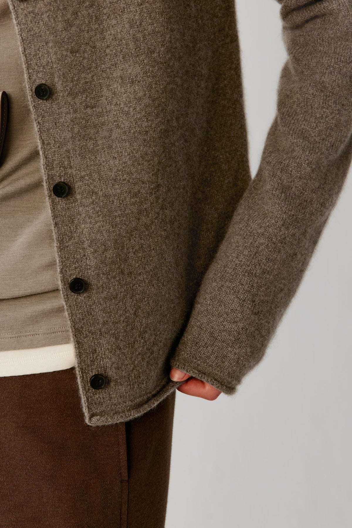 Close-up of a person wearing a taupe-brown knit button-front cardigan over a light beige top and brown trousers, with the hand holding the hem.