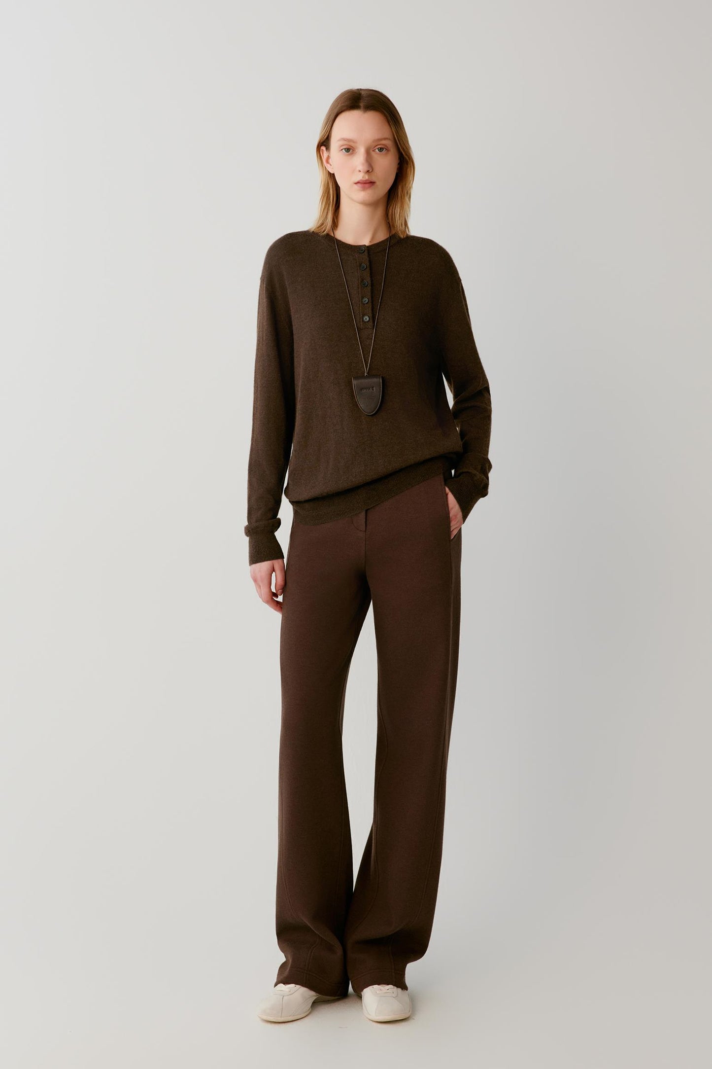 A model stands against a light gray studio background wearing a dark brown long-sleeve knit top and matching wide-leg knit pants, with a long pendant necklace and off-white slip-on shoes.