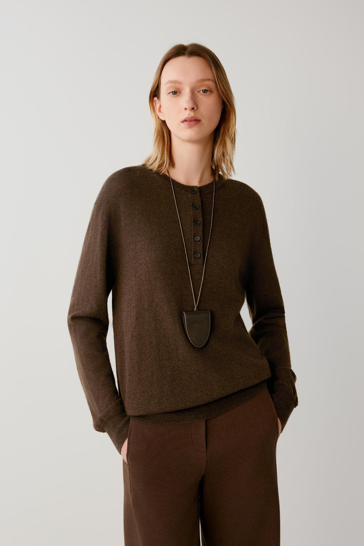 A woman modeling a dark brown long-sleeve knit sweater with a buttoned henley neckline, paired with matching brown trousers and a long dark pendant necklace, against a plain light gray studio background.