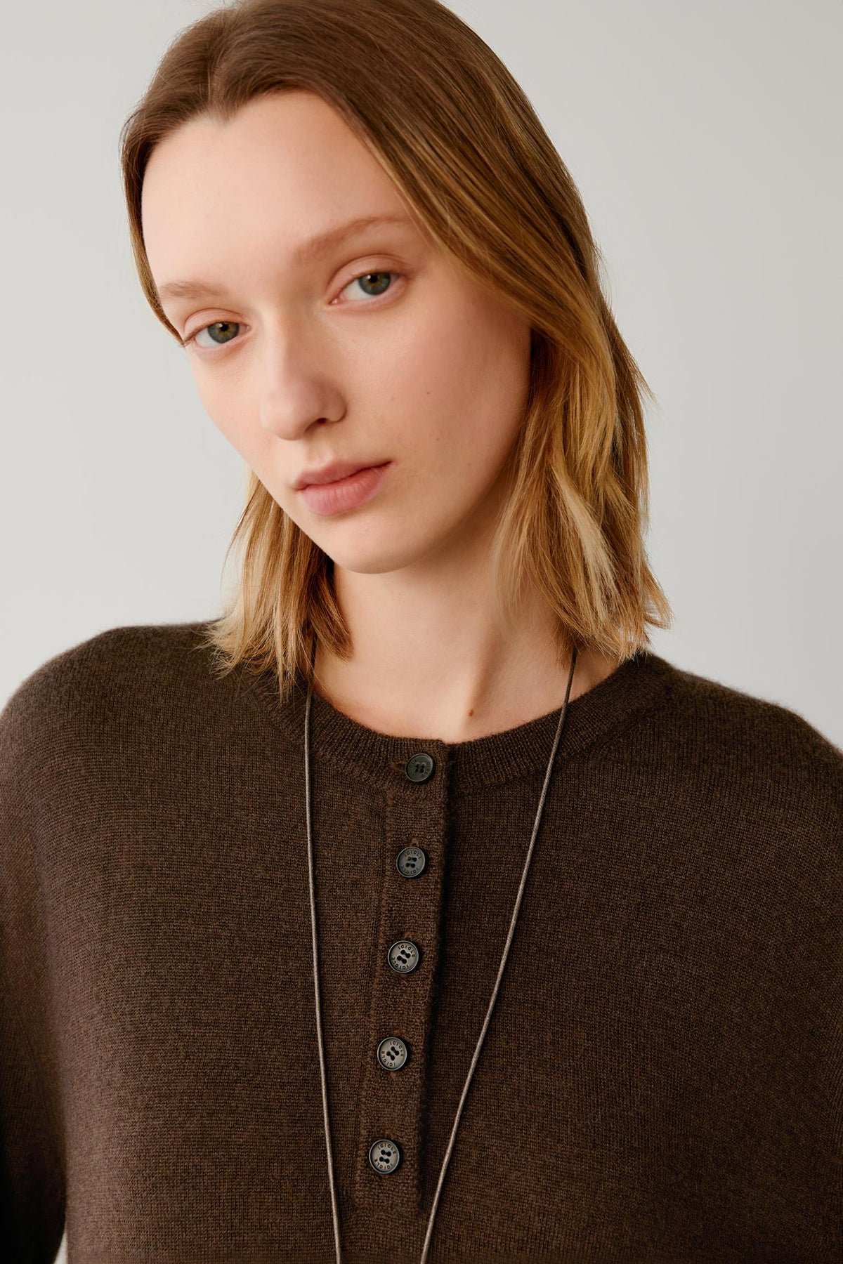 A close-up shot of a woman wearing a dark brown knit sweater with a buttoned henley-style front and a long black cord necklace, set against a plain light gray background.