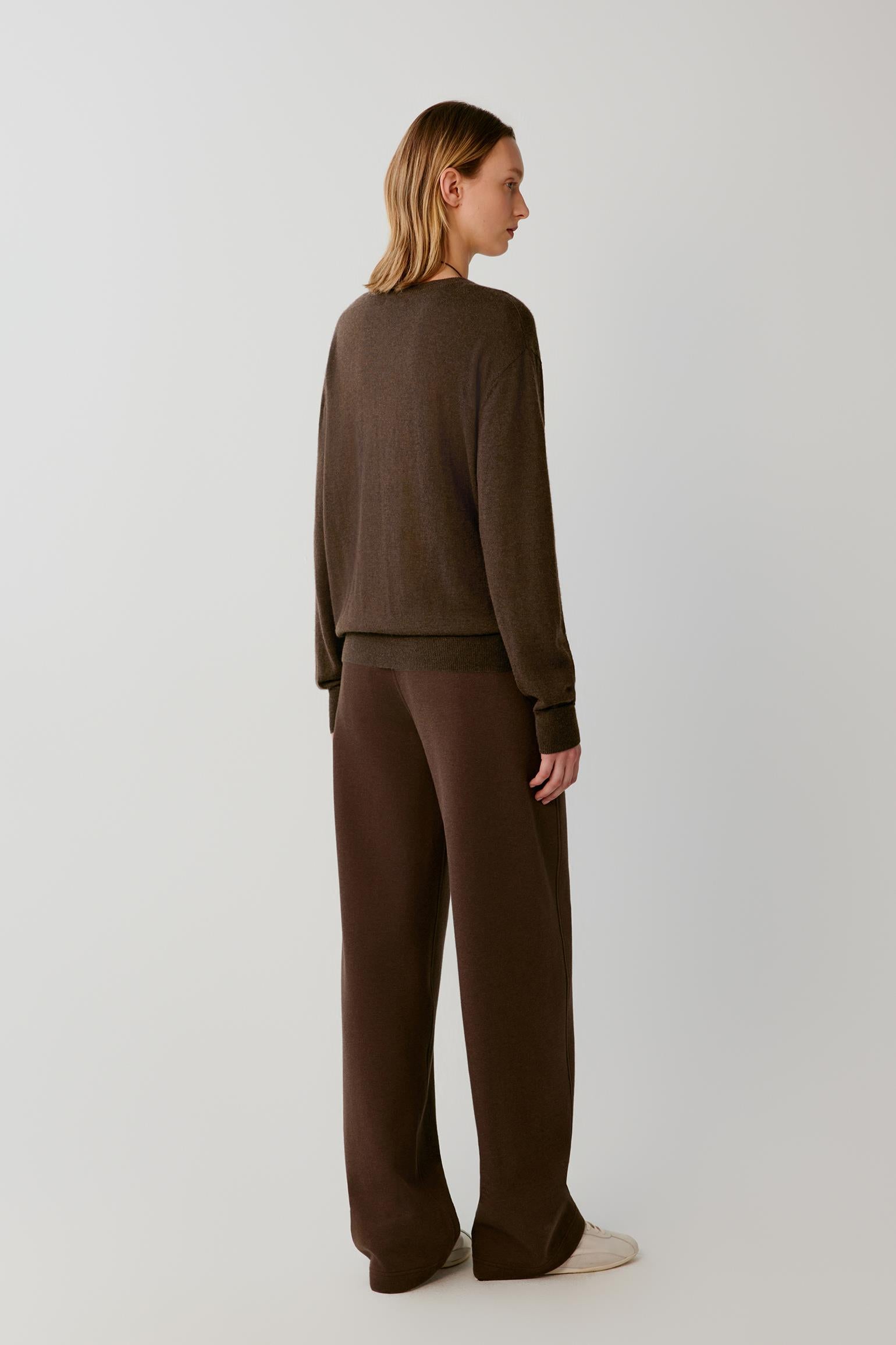 A woman stands in profile wearing a dark brown knit long-sleeve top and matching wide-leg pants against a plain light gray studio background.