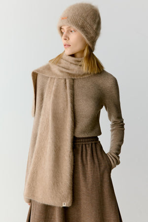 Portrait view of a woman wearing a taupe cashmere scarf wrapped around the neck and draped over the shoulders. The image highlights the plush texture, soft volume and fluid drape of the cashmere, while the tonal styling emphasizes the warm, understated character of the accessory.