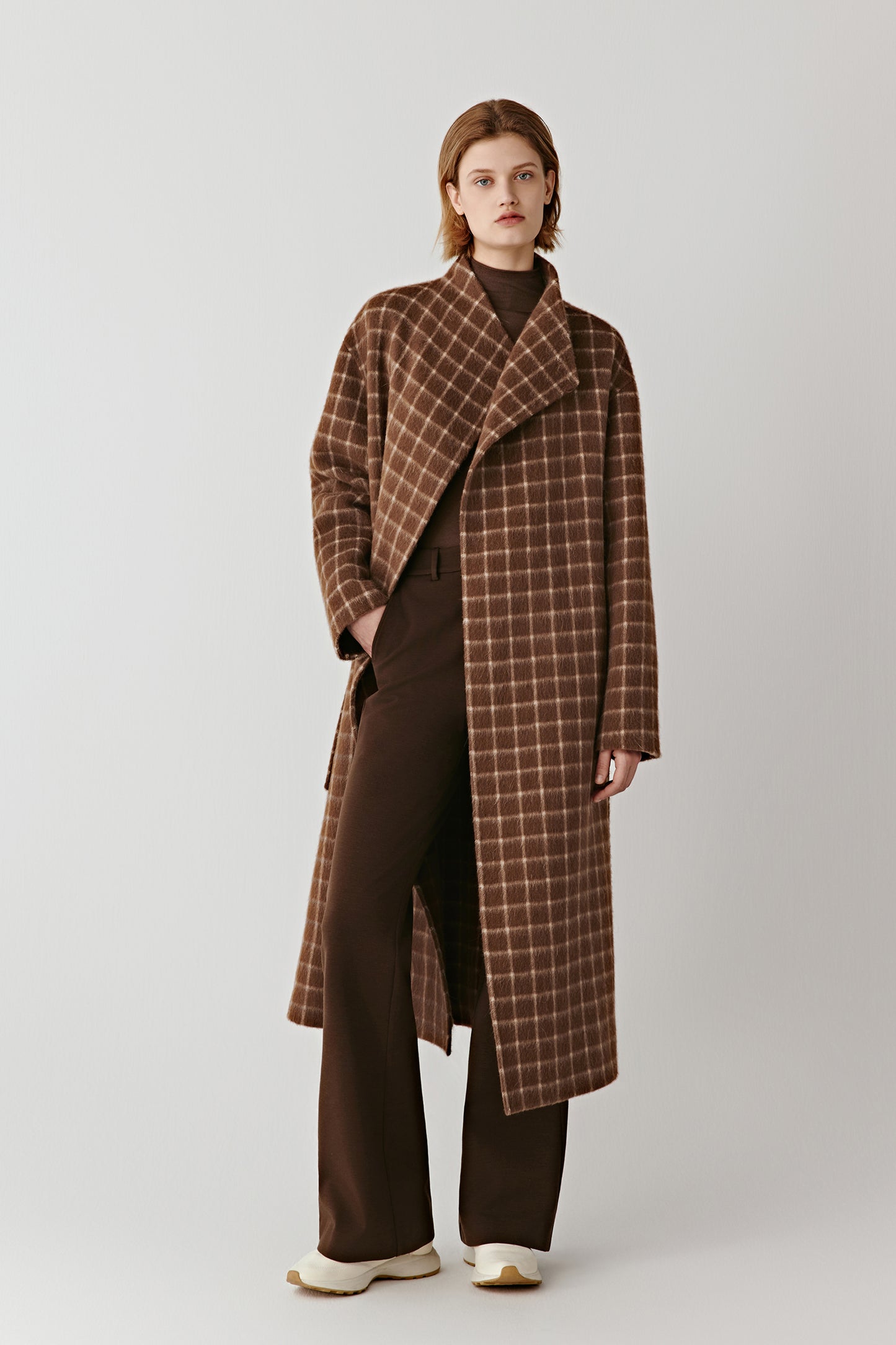 Full-length front view of a person wearing a long double-faced checked wool coat in brown tones, styled with a relaxed, timeless silhouette.
