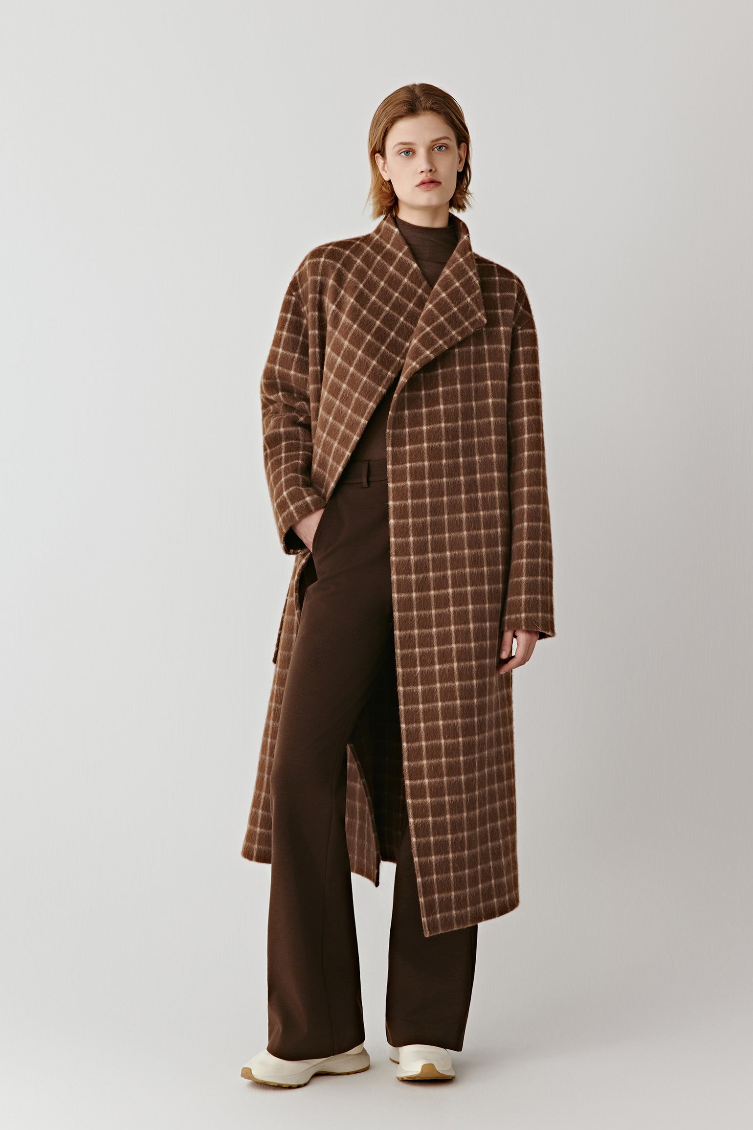 Full-length front view of a person wearing a long double-faced checked wool coat in brown tones, styled with a relaxed, timeless silhouette.