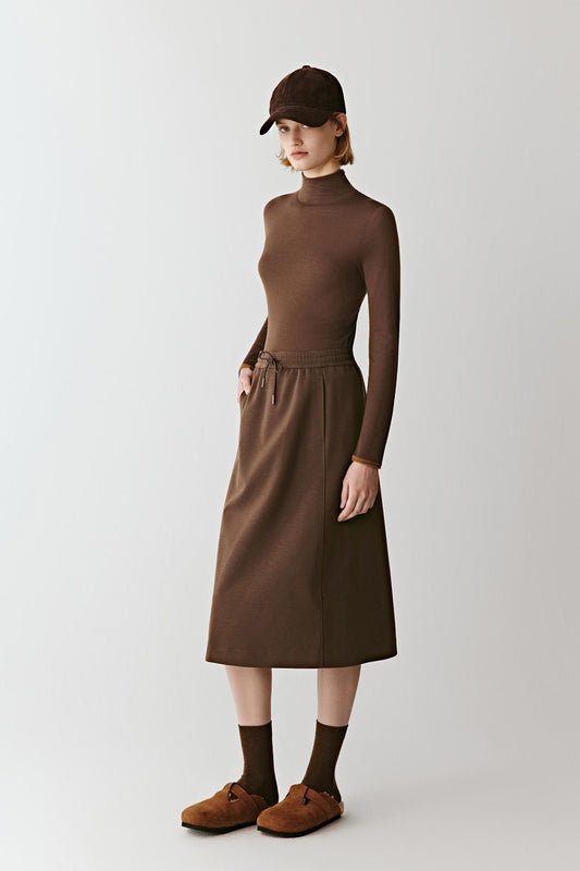 Full-length view of a person wearing a fitted mock-neck pullover dress in deep red-brown merino wool, standing against a light background.