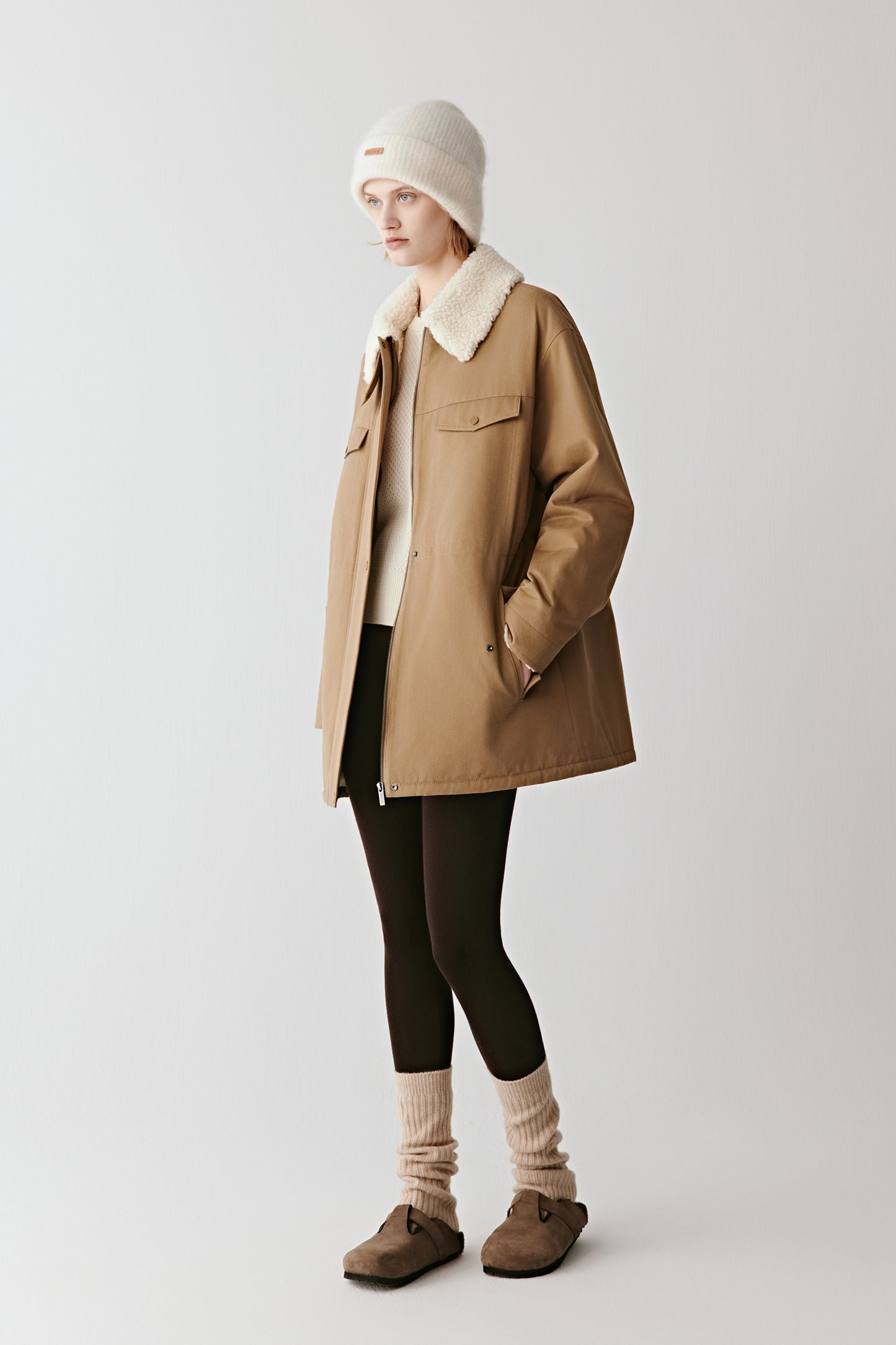 Full-length front view of a person wearing a deep camel cotton twill parka with a shearling-effect collar, paired with dark trousers and winter boots.