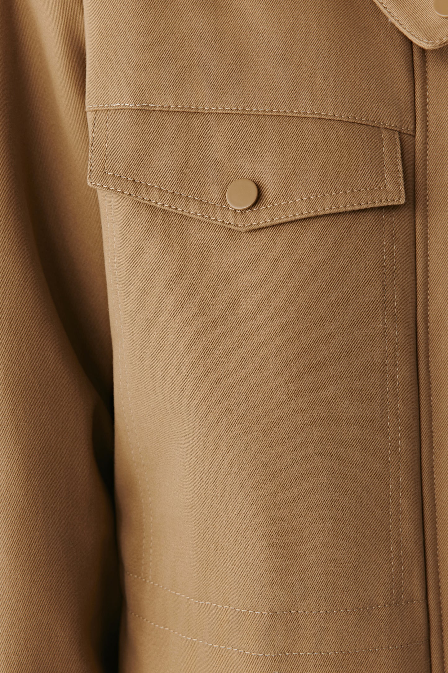 Detail close-up of the camel cotton twill parka showing a flap pocket and the fine twill texture of the fabric.