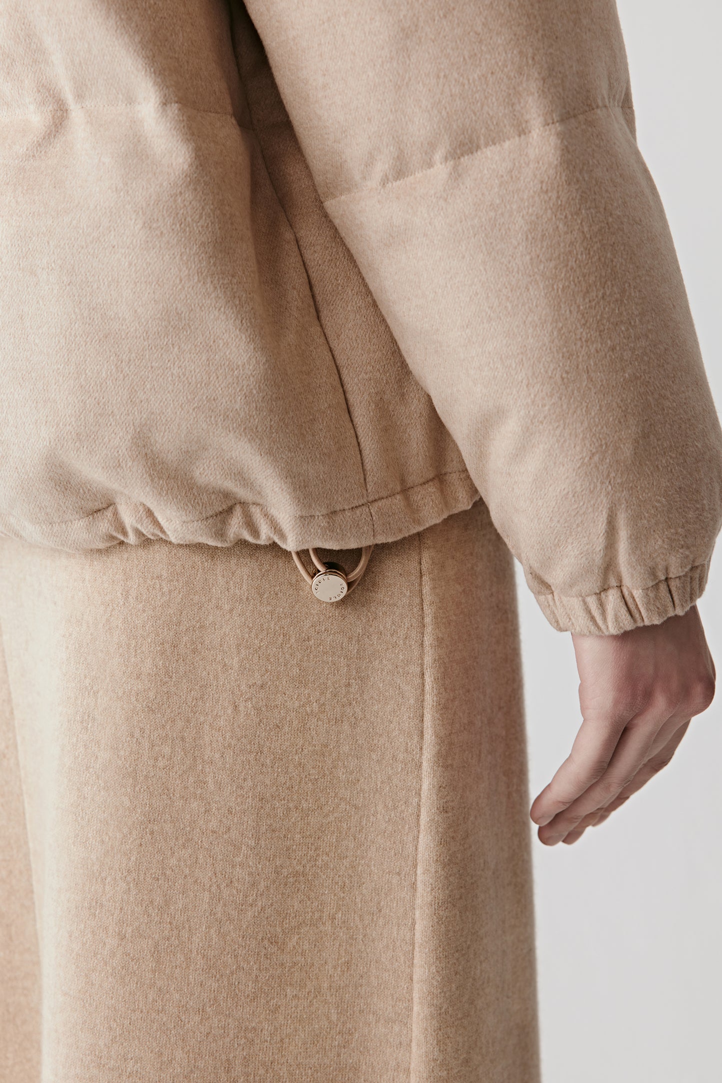Detail close-up of the camel down coat’s adjustable hem with a discreet metal toggle and soft tweed fabric.