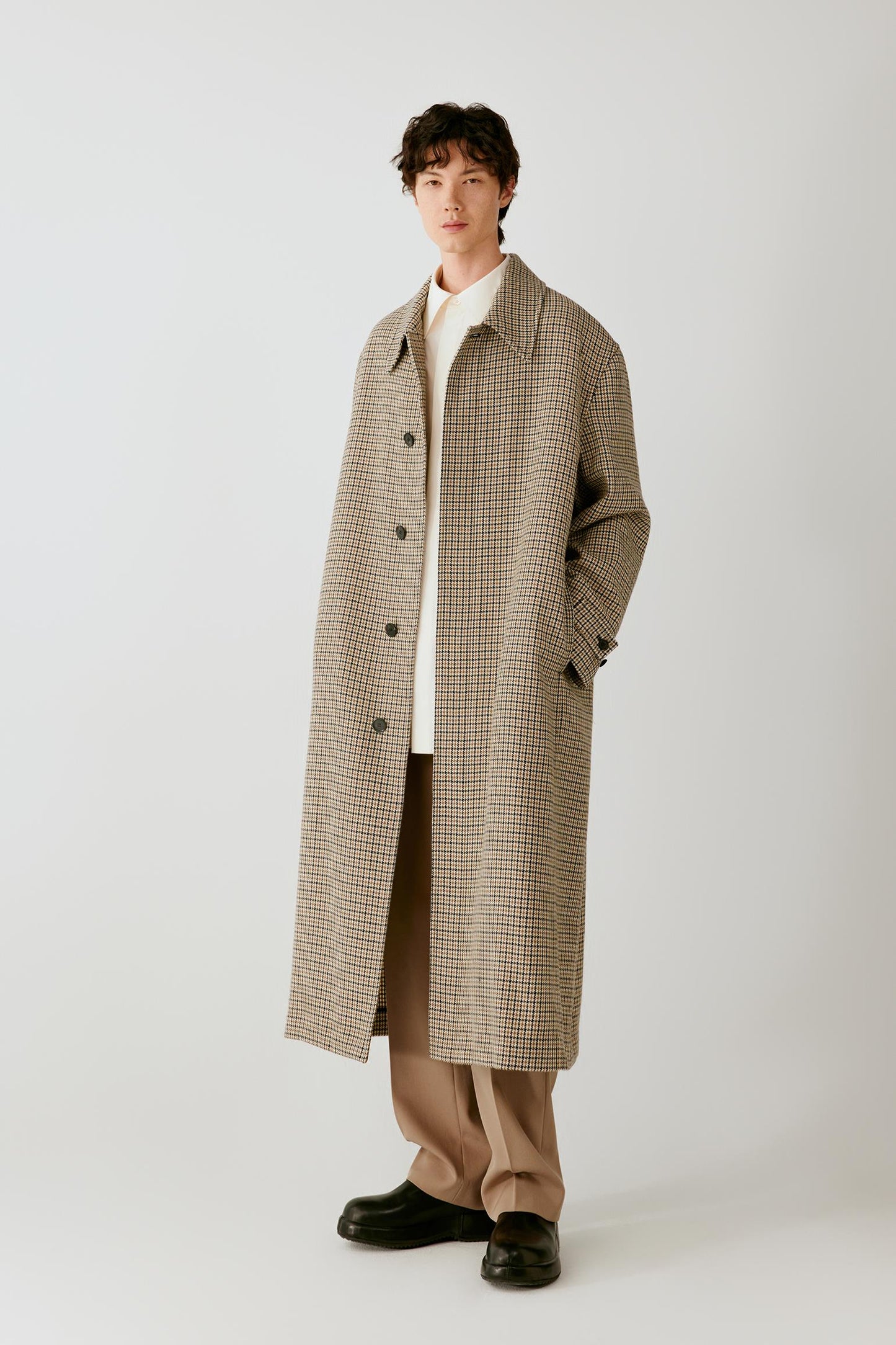Front view of a man wearing a beige and black houndstooth wool coat, highlighting the long silhouette, pointed collar and concealed fastening.