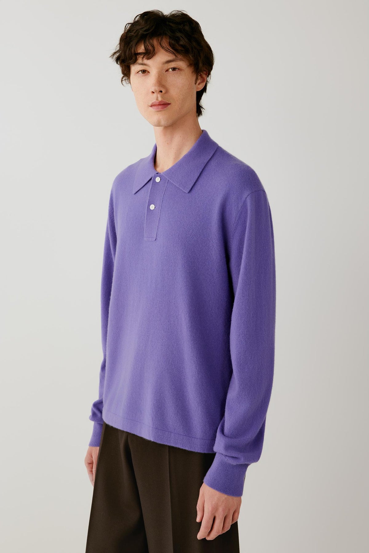 A model stands against a light gray studio background wearing a purple long-sleeve polo knit top and dark brown wide-leg trousers.