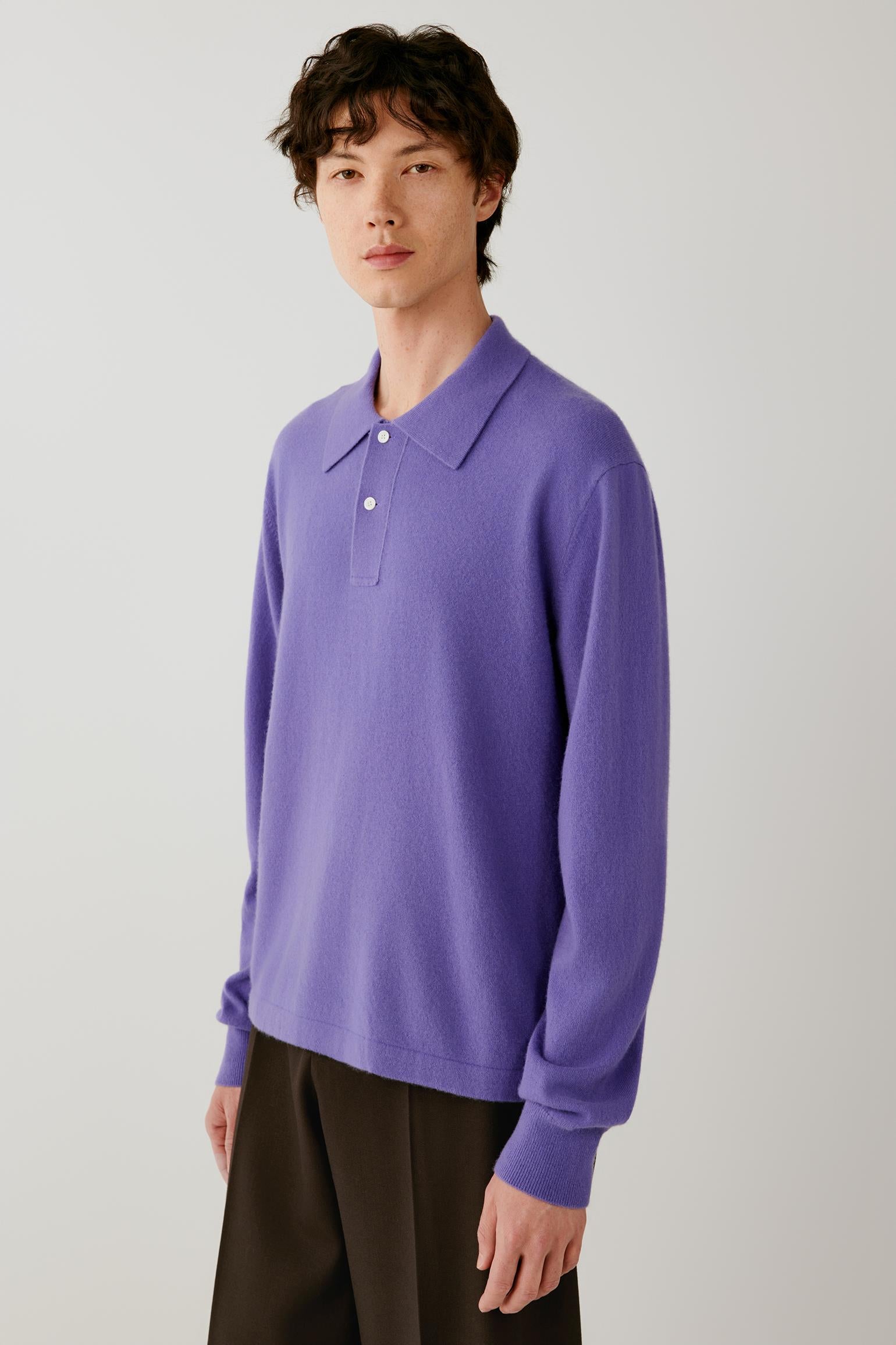 A model stands against a light gray studio background wearing a purple long-sleeve polo knit top and dark brown wide-leg trousers.