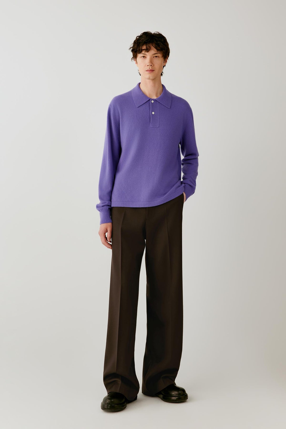 A model stands against a light gray studio background wearing a purple long-sleeve polo knit top and dark brown wide-leg trousers.