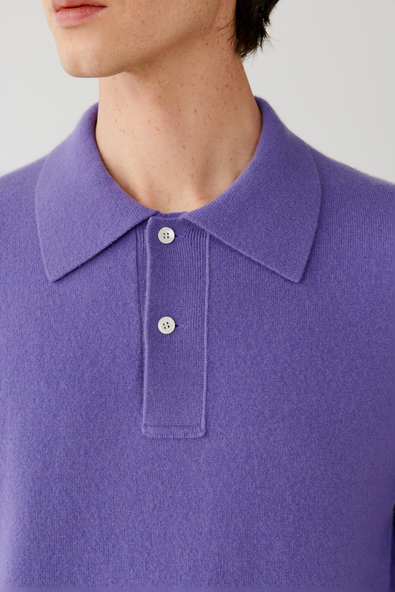 Close-up of a person wearing a purple knit polo sweater with a collar and two white buttons, against a light neutral background.