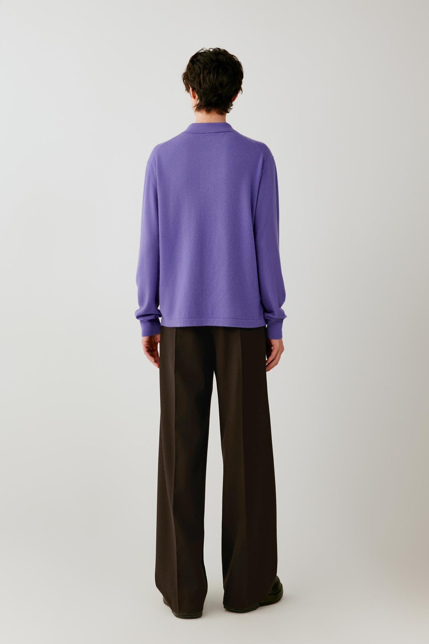 A model stands from the back against a light gray studio background wearing a purple long-sleeve polo knit top and dark brown wide-leg trousers.
