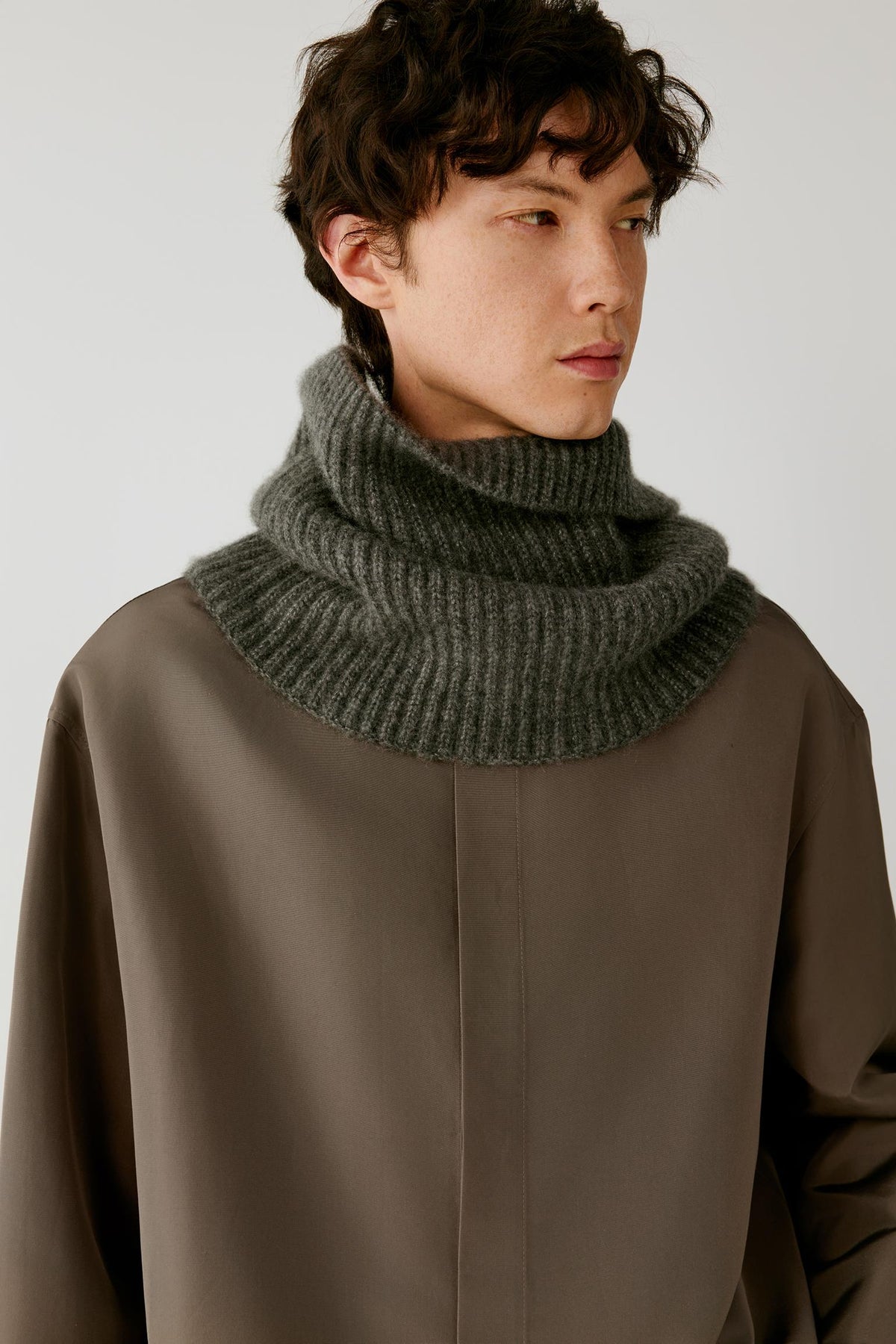 A man wearing a dark gray ribbed knit cowl neck scarf and a brown jacket, photographed against a neutral light gray background.