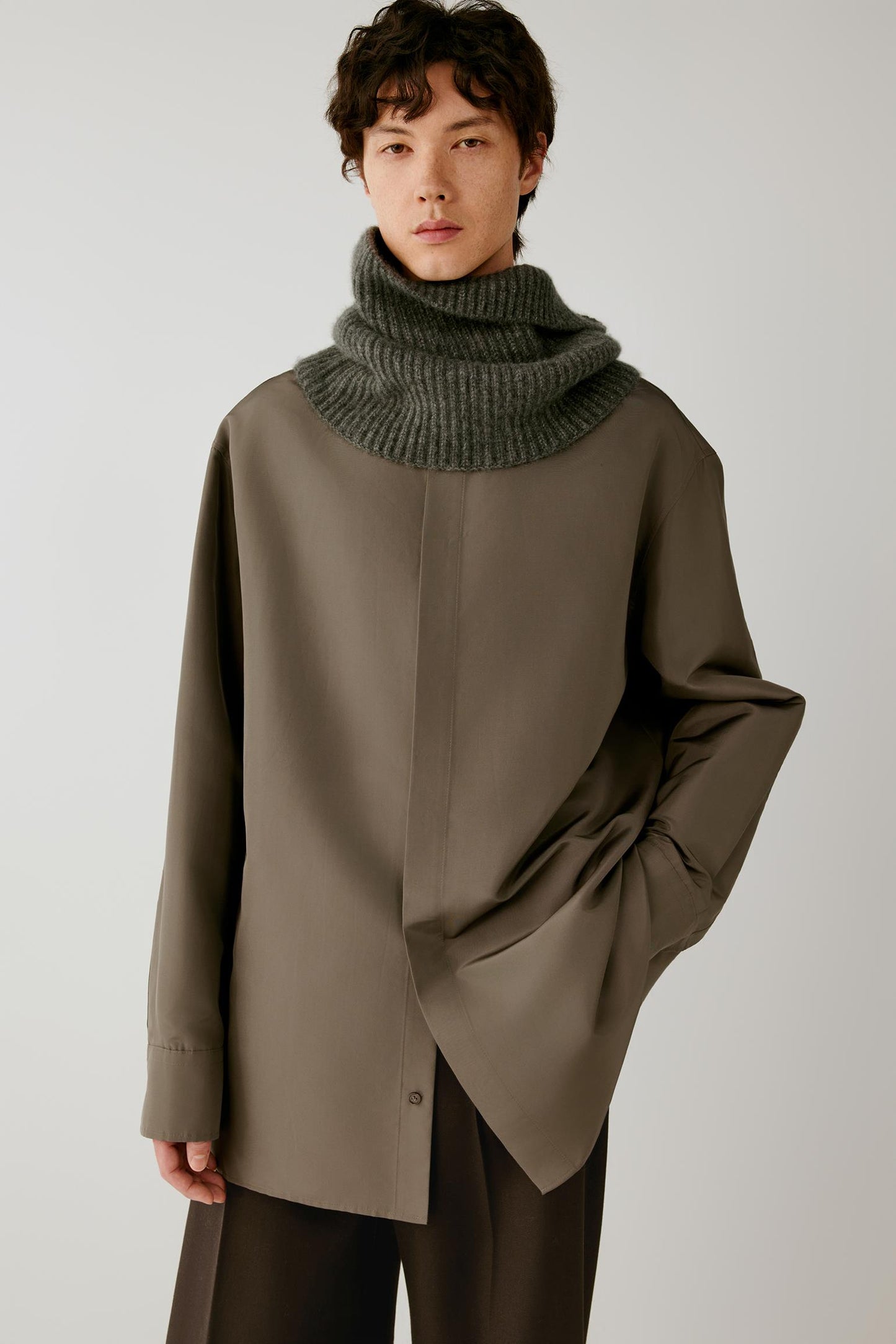 A model wearing an oversized taupe-brown asymmetrical button-up shirt with a chunky dark green ribbed knit cowl neck scarf, set against a plain light gray studio background.