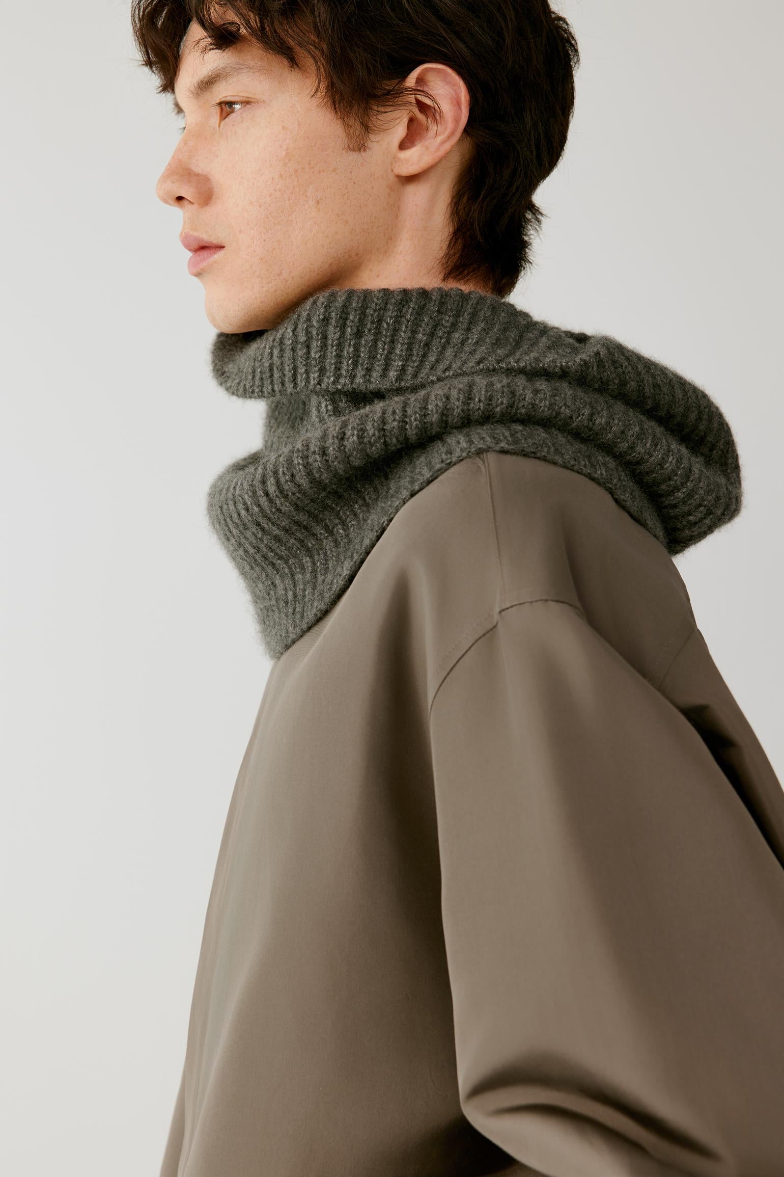 A close-up side profile of a model wearing a thick, ribbed knit cowl neck scarf in dark gray, paired with an oversized taupe-brown pullover sweater against a light gray studio background.