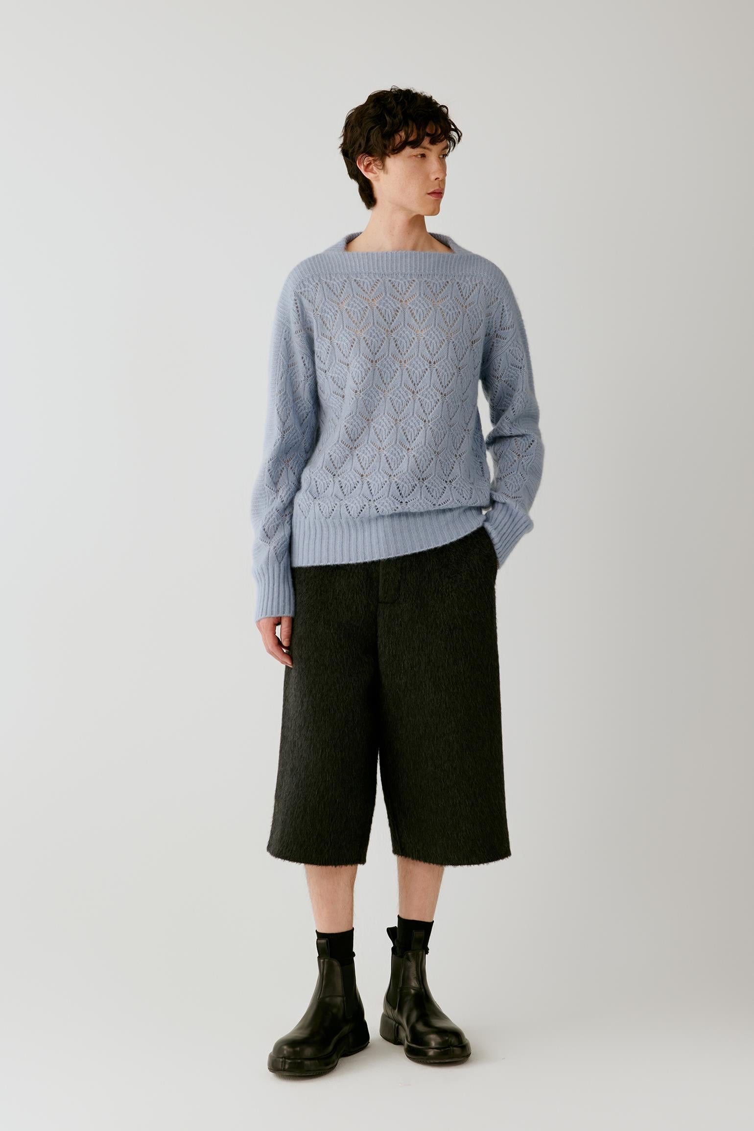A model stands against a light gray studio background wearing a light blue cable-knit sweater, dark charcoal cropped trousers, black socks, and black boots.